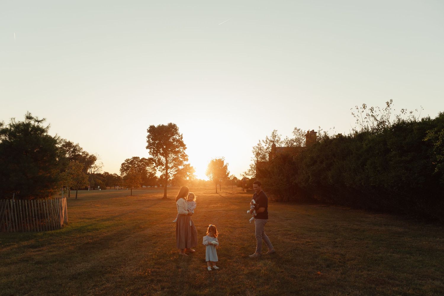 Family silhouetted against golden sunset in open field with trees and wooden fence in background.