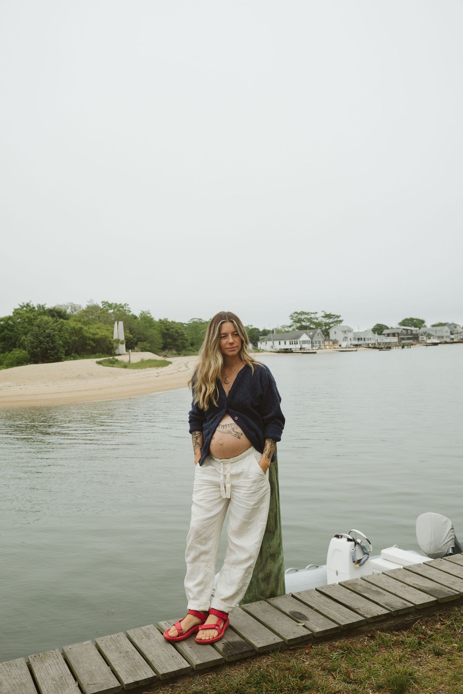 Pregnant woman in navy top and white pants stands on a wooden dock by calm water on an overcast day.
