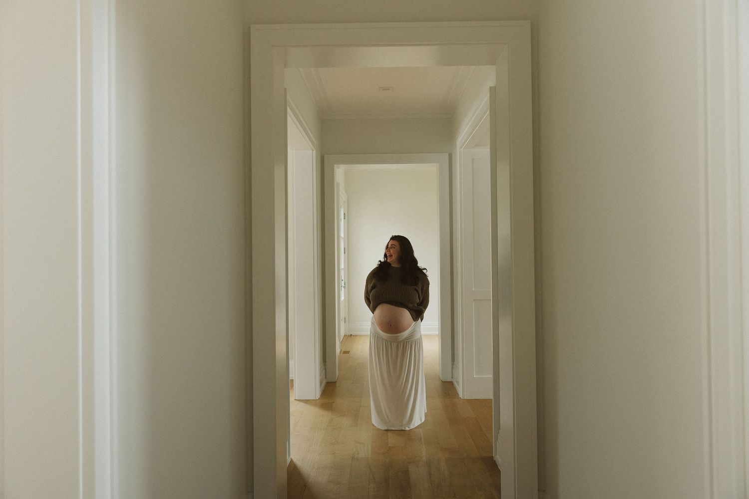 Pregnant woman in white skirt stands in a bright white hallway, cradling her baby bump in a serene maternity photo.