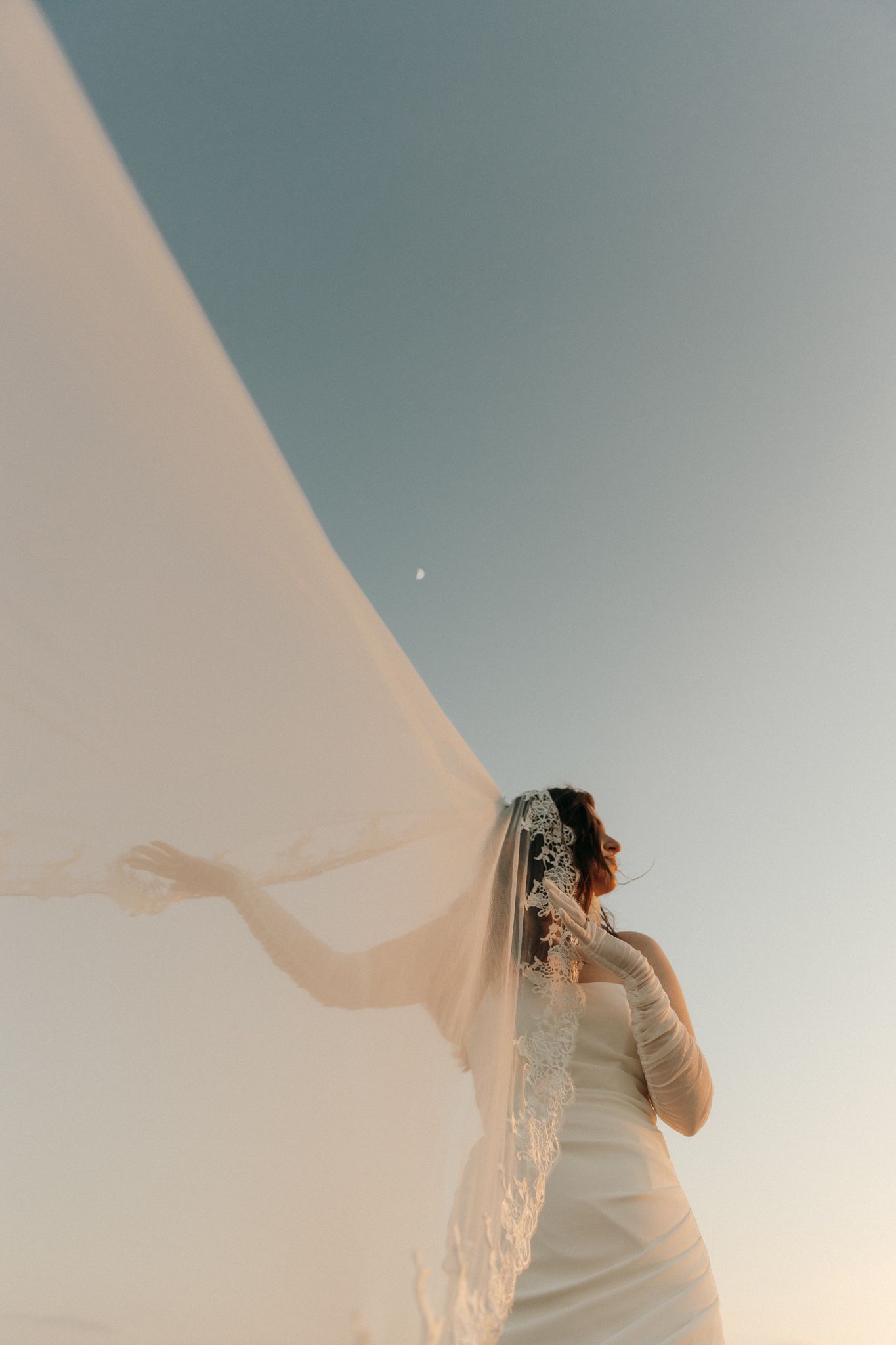 Bride in white gown holds flowing lace veil against blue sky with crescent moon at golden hour.