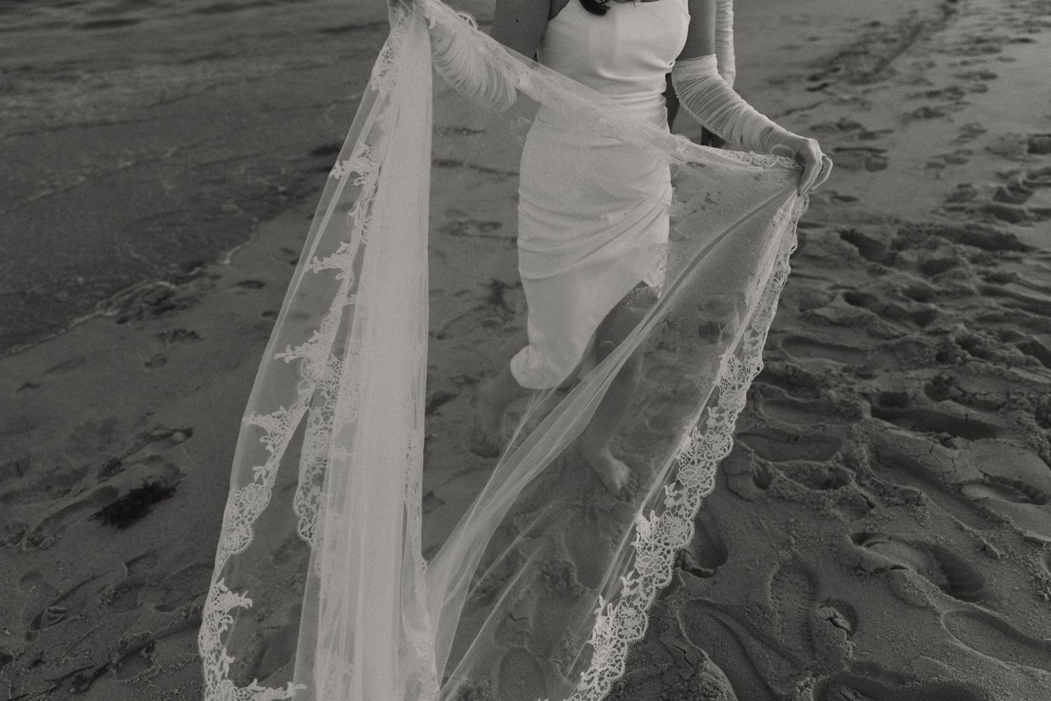 Black and white beach wedding photo of bride in fitted gown holding lace veil train on sandy shore.