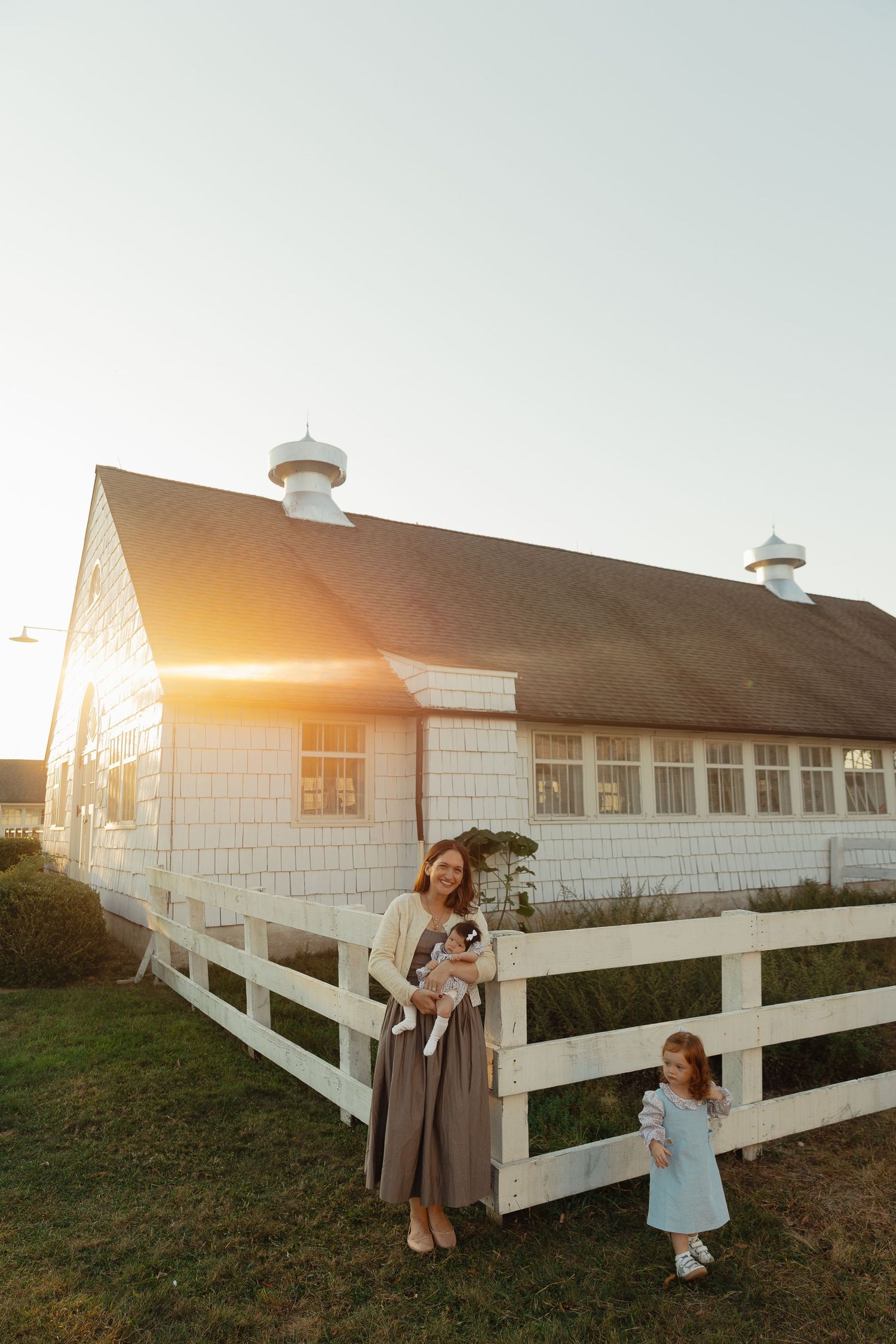 Woman holding baby and young girl standing by white fence and barn at golden hour sunset on a farm.