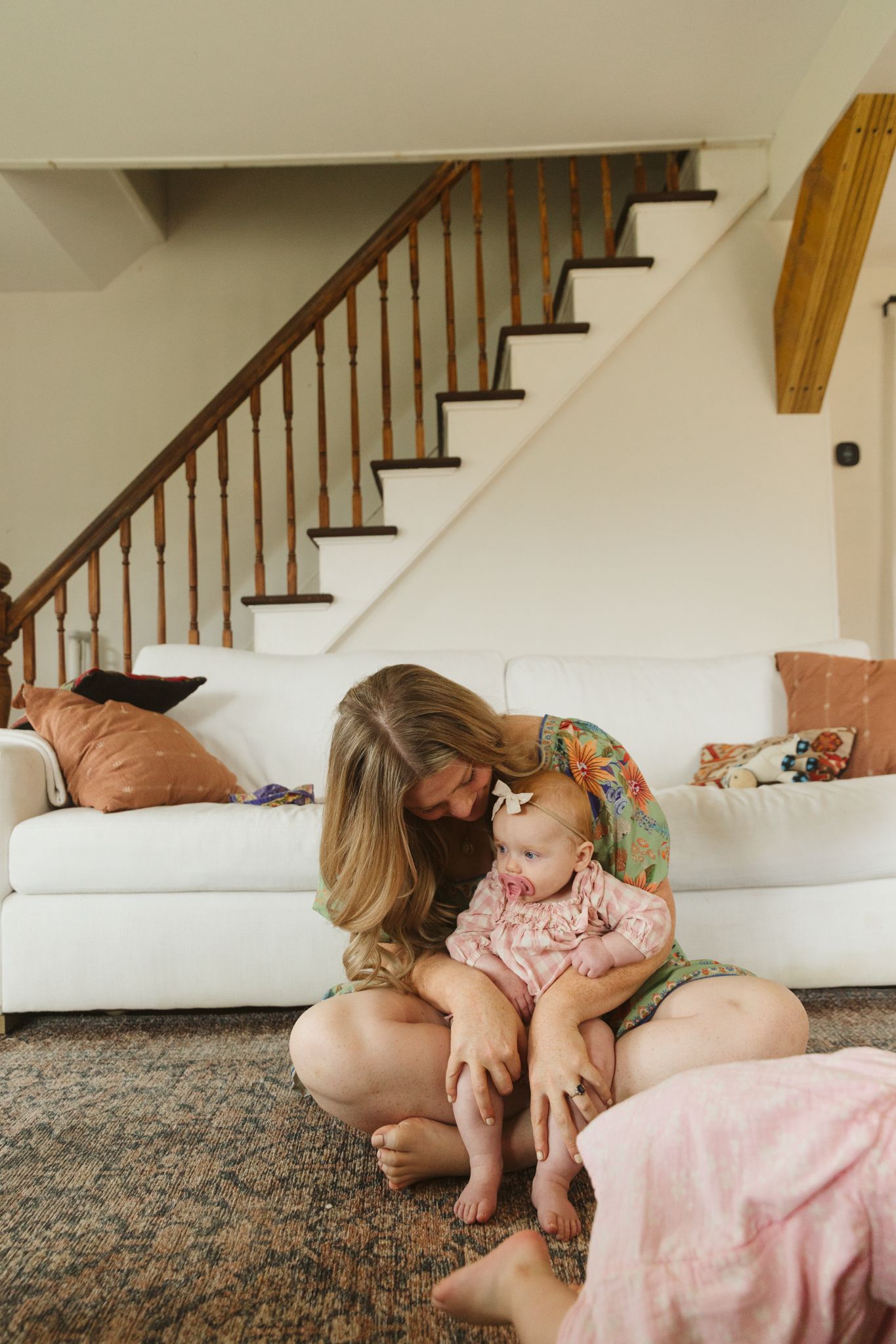 Loving mother sitting on floor cradling baby girl in floral onesie near white couch with staircase.