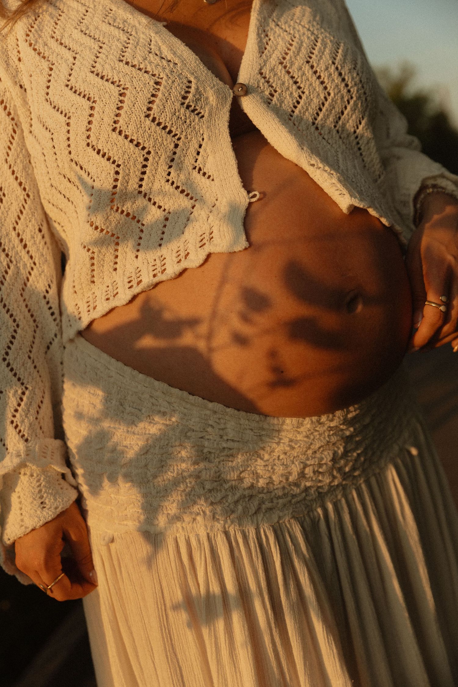 Woman in white crochet crop top and pleated skirt with golden sunlight and leaf shadows on her midriff.