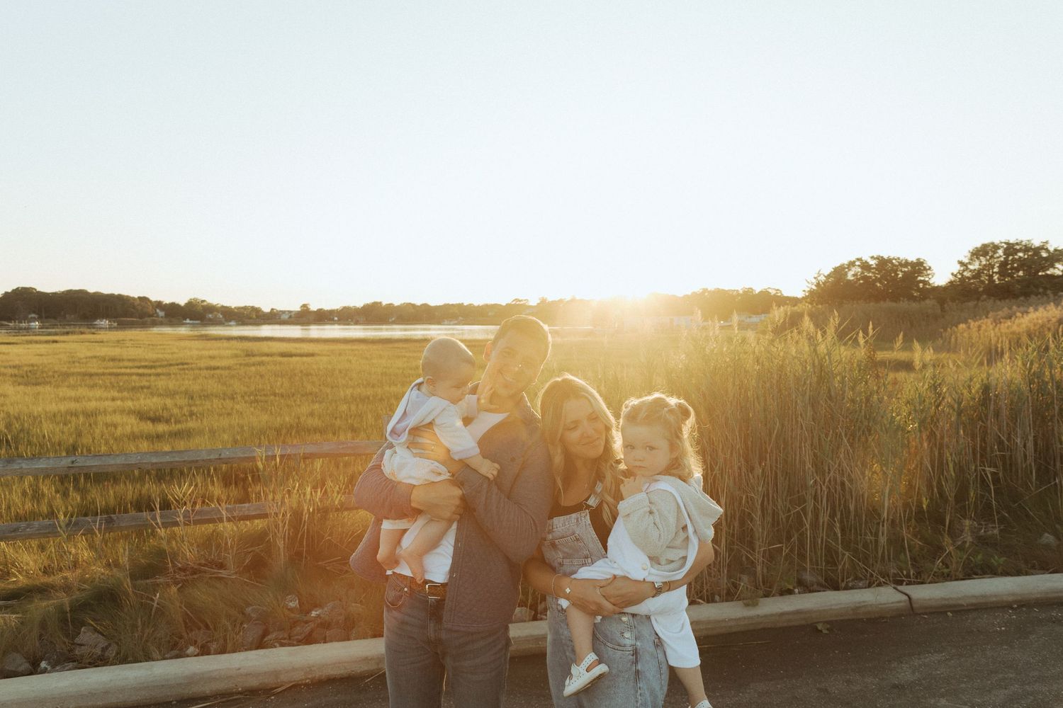 Family of four embracing at golden hour in a sunny field, backlit by warm sunset light.