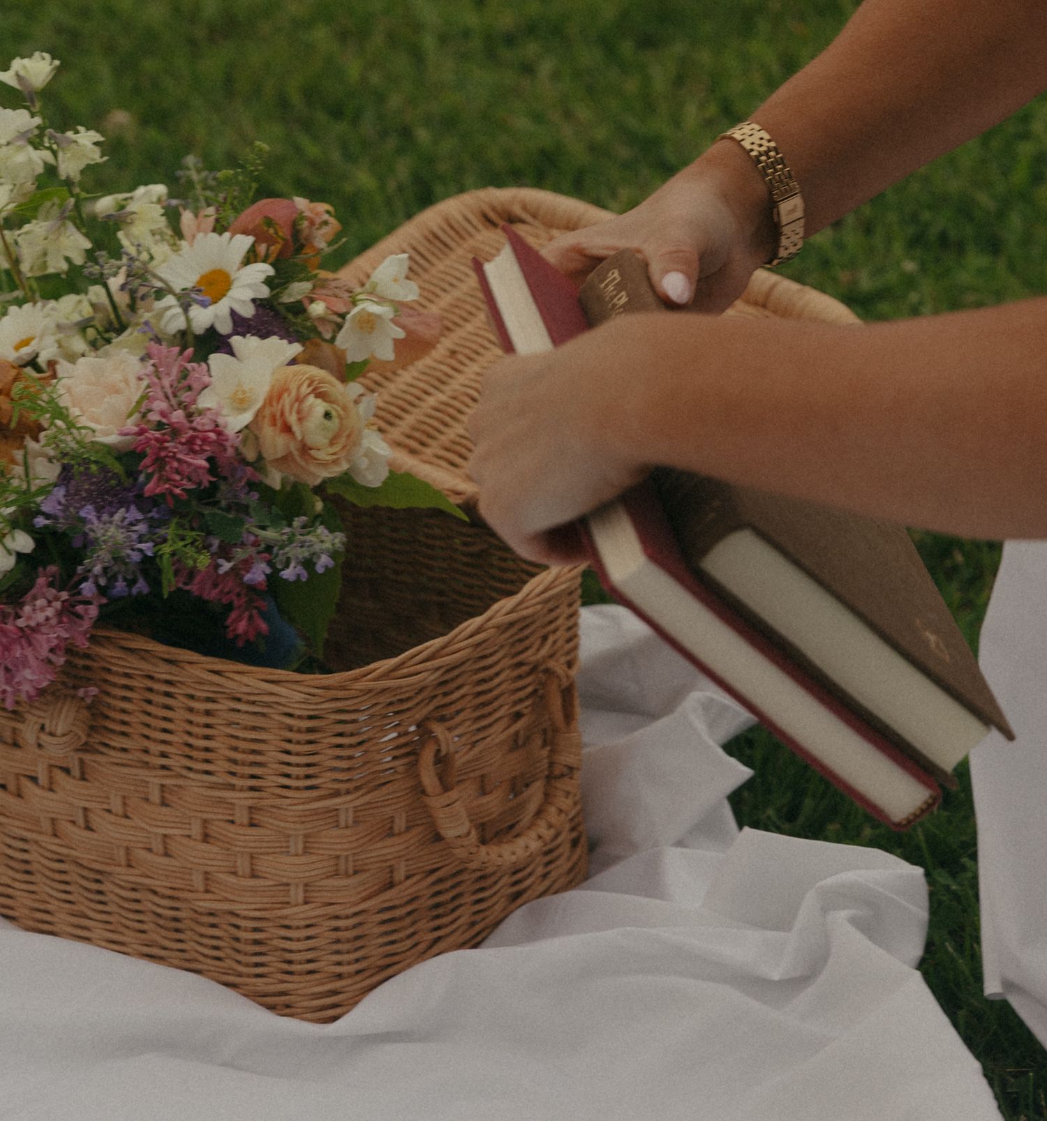 Person placing books into a wicker picnic basket filled with fresh wildflowers on a white blanket in green grass.
