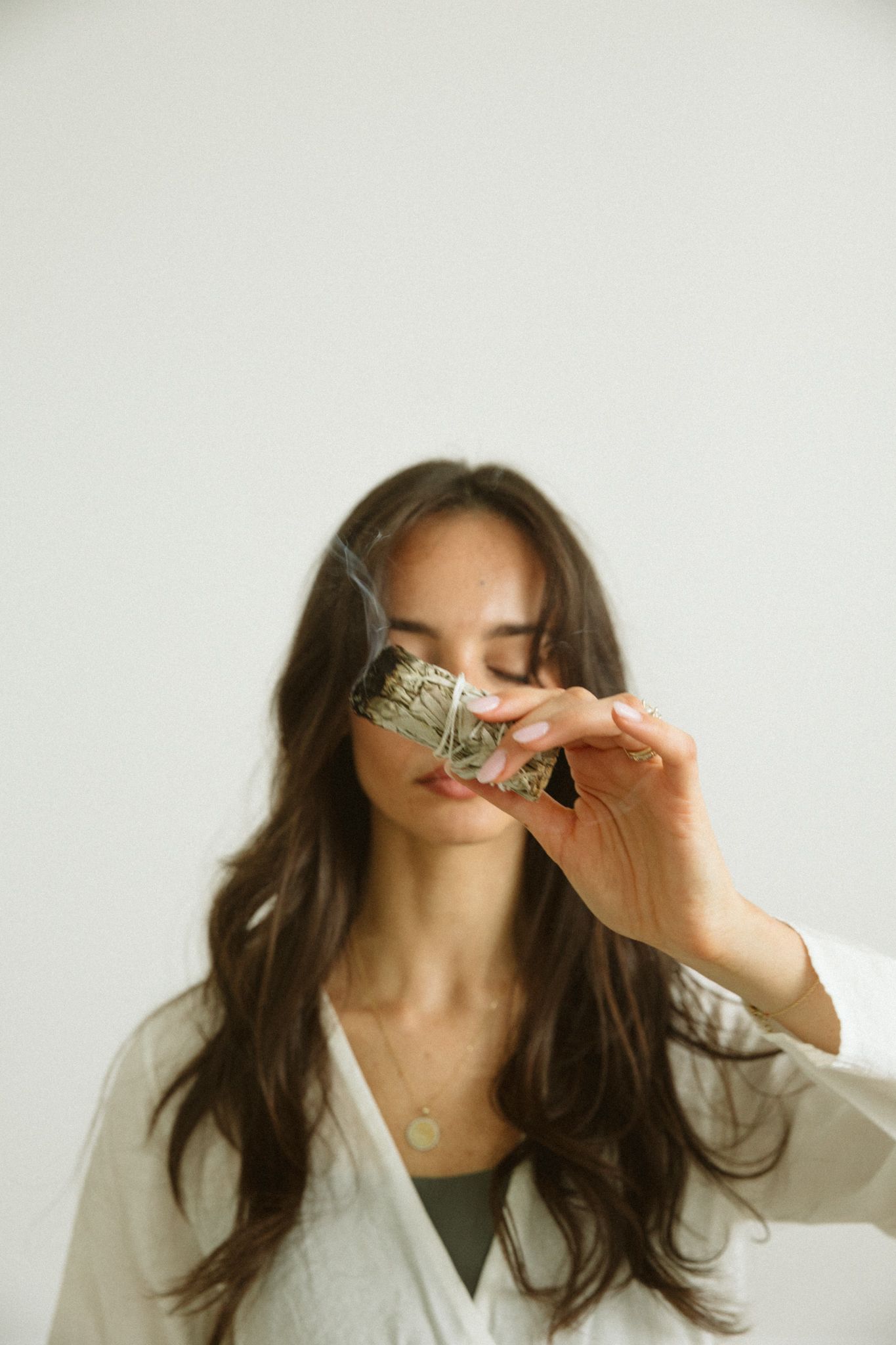 Woman with long dark hair holding a smoking sage smudge stick near her face, wearing a white robe.