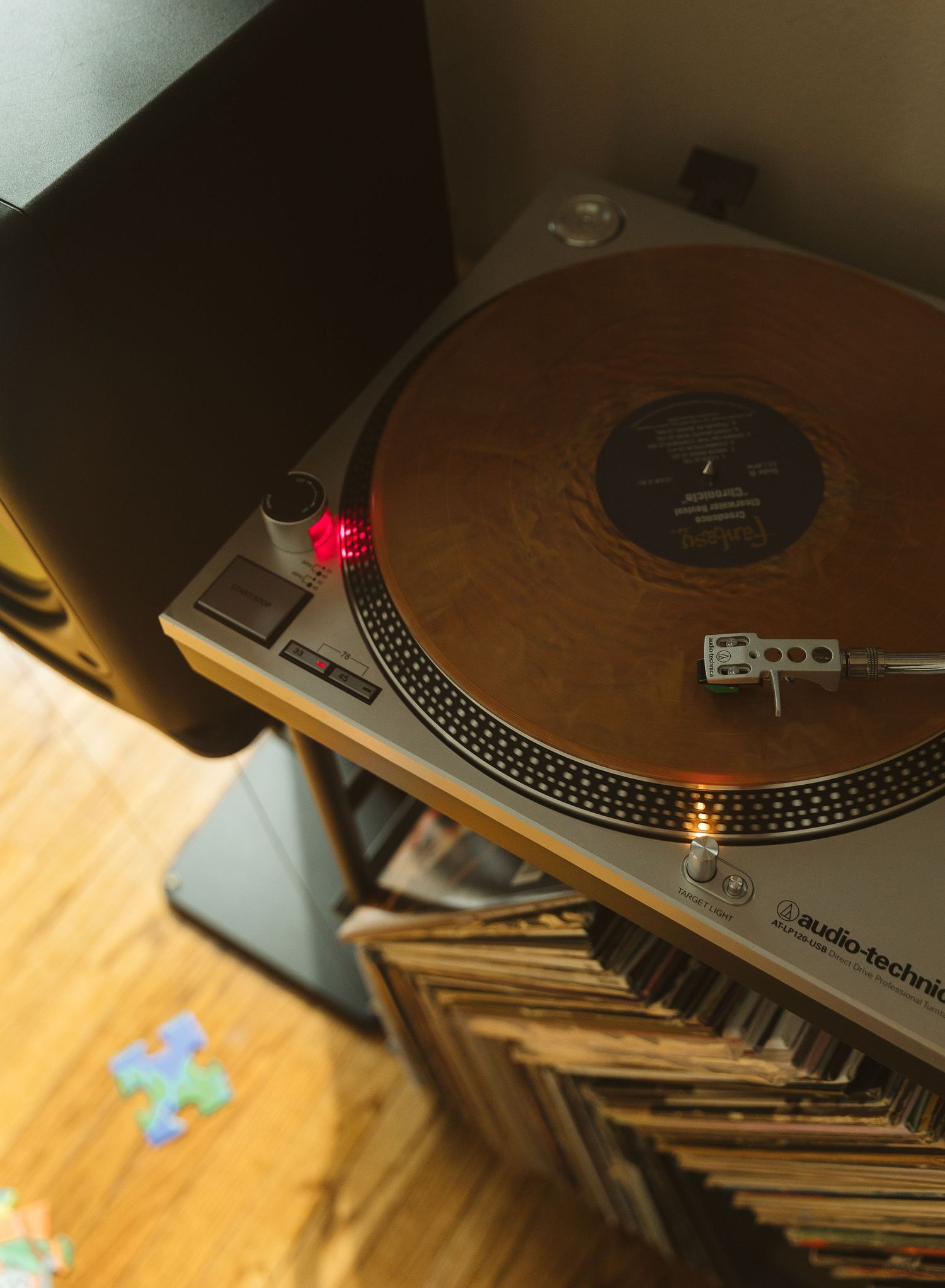 Audio-Technica turntable with golden vinyl record spinning, surrounded by stacked album collection on wooden floor.