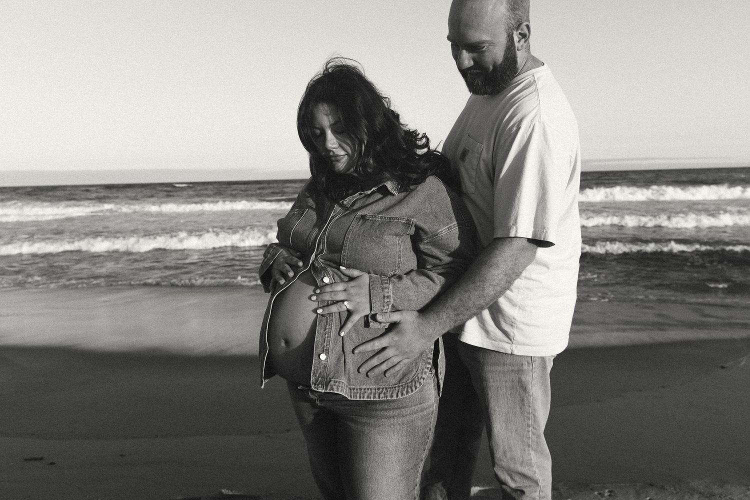 Black and white beach photo of a pregnant woman and man posing together by the ocean waves.