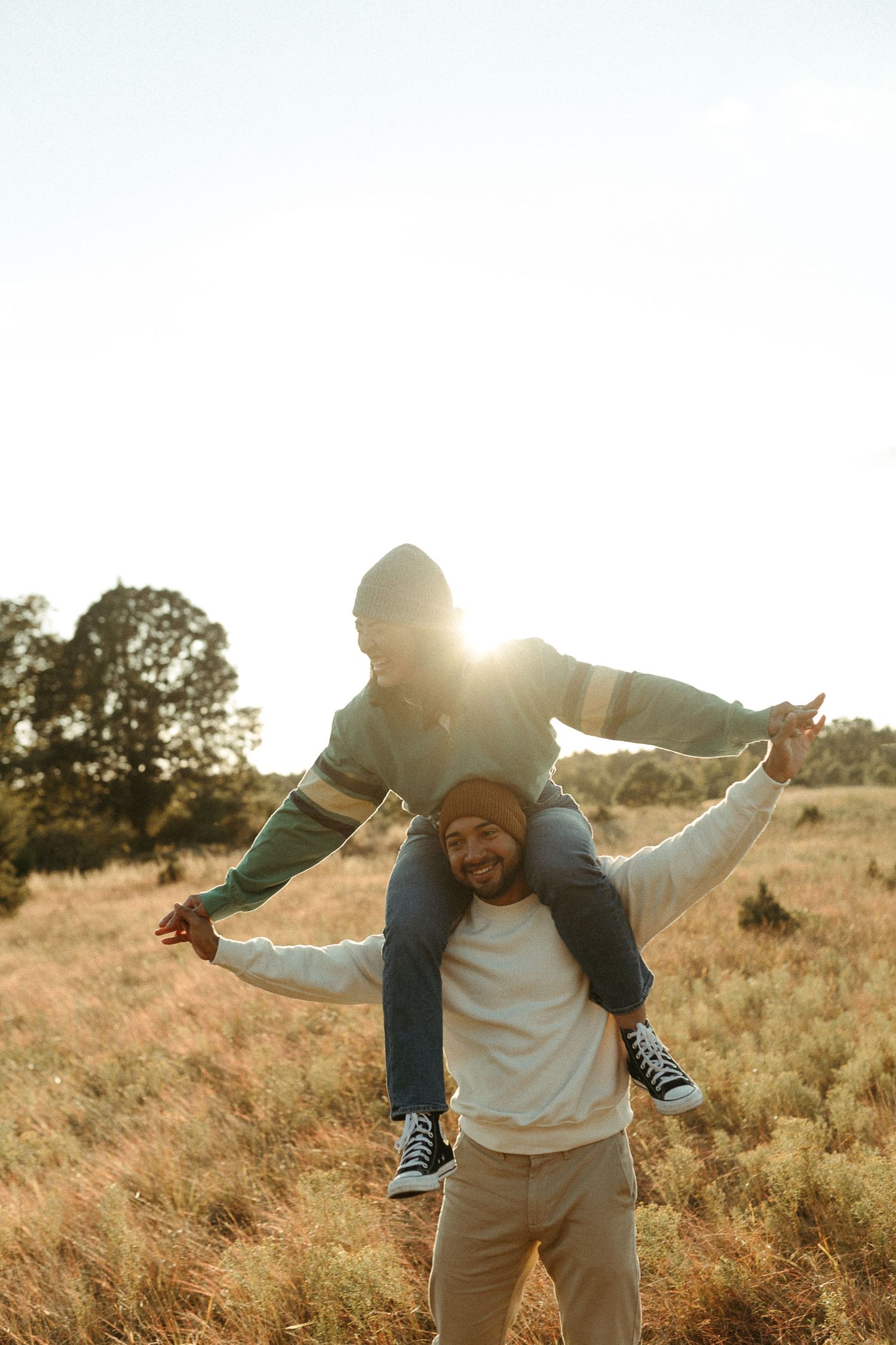 Smiling father carries child on shoulders with arms outstretched in sunny golden field.