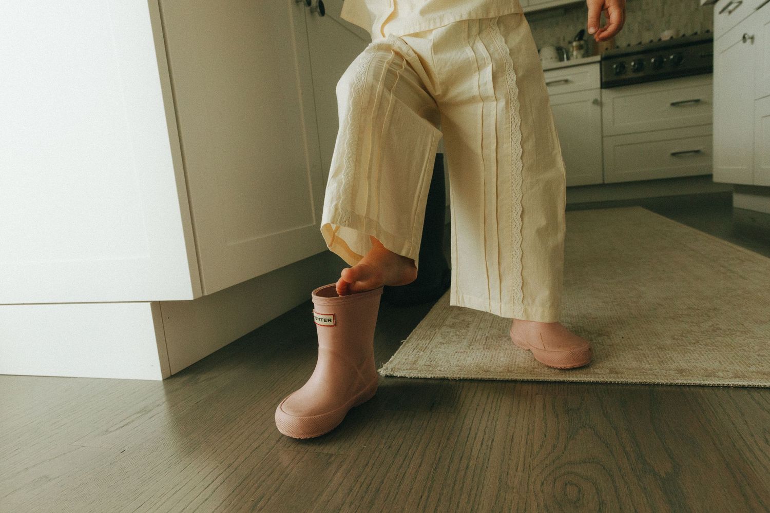 Young child wearing one pink rain boot stands on kitchen floor in cream wide-leg pants.