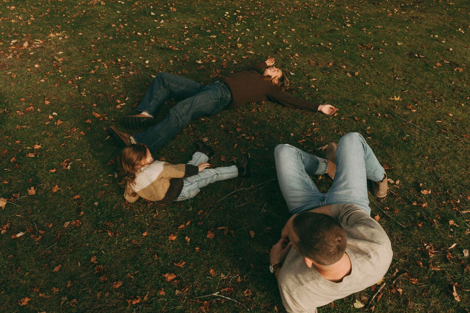 People laying together on grass covered in fallen autumn leaves.