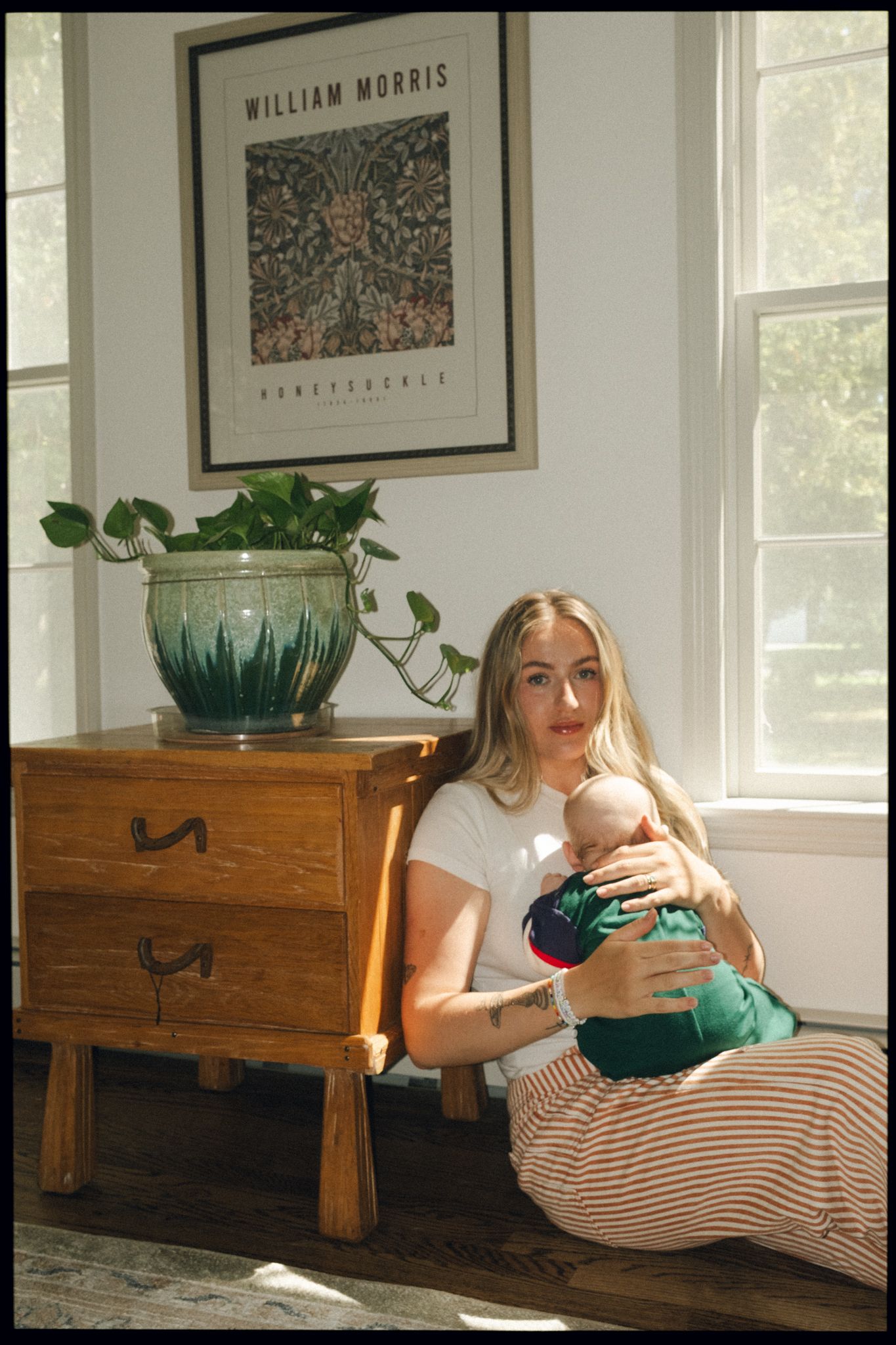 Young mother sitting on floor holding newborn baby beside wooden dresser with plant under William Morris art print.