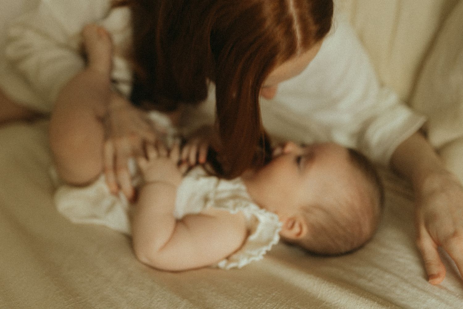 A mother tenderly kisses her newborn baby lying on a soft white blanket in a warm, intimate setting.