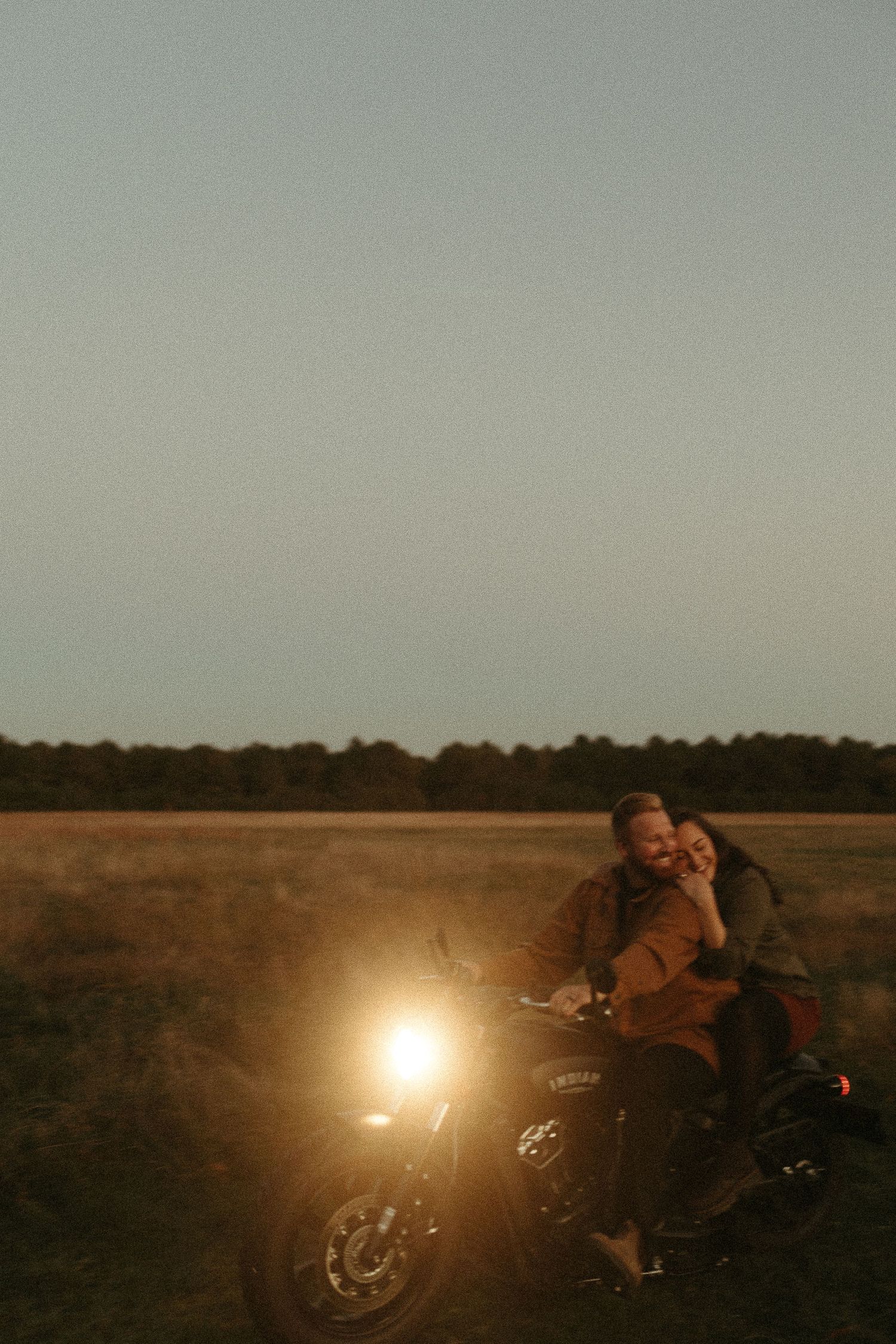 Couple sitting together on a motorcycle with headlight glowing in an open field at dusk.