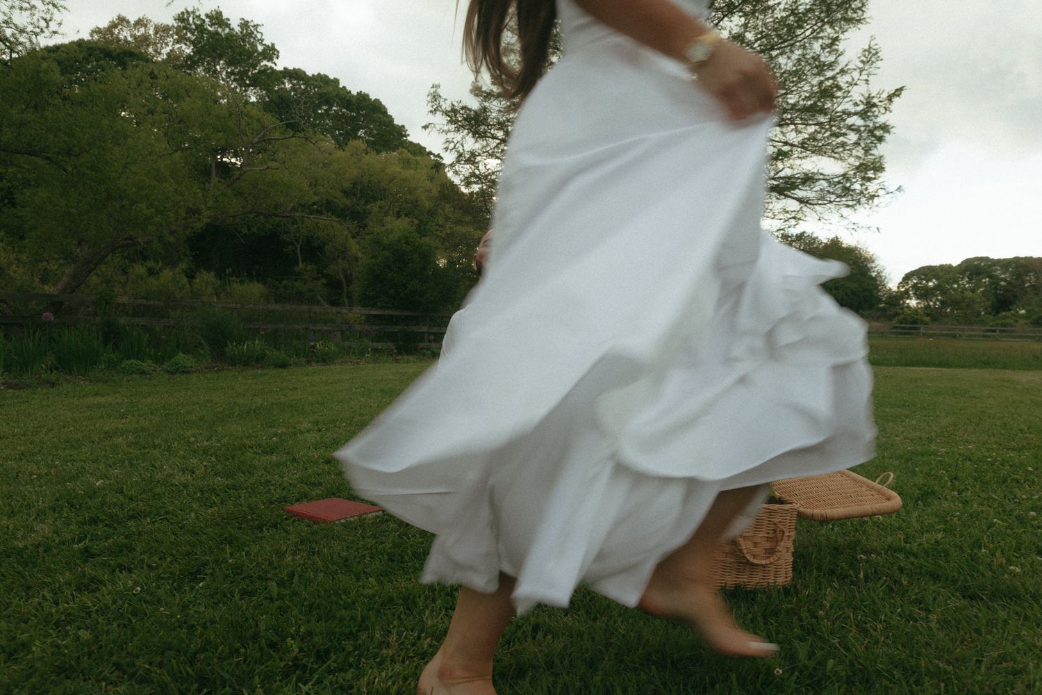 A woman in a flowing white dress twirls barefoot on a lush green lawn surrounded by trees.