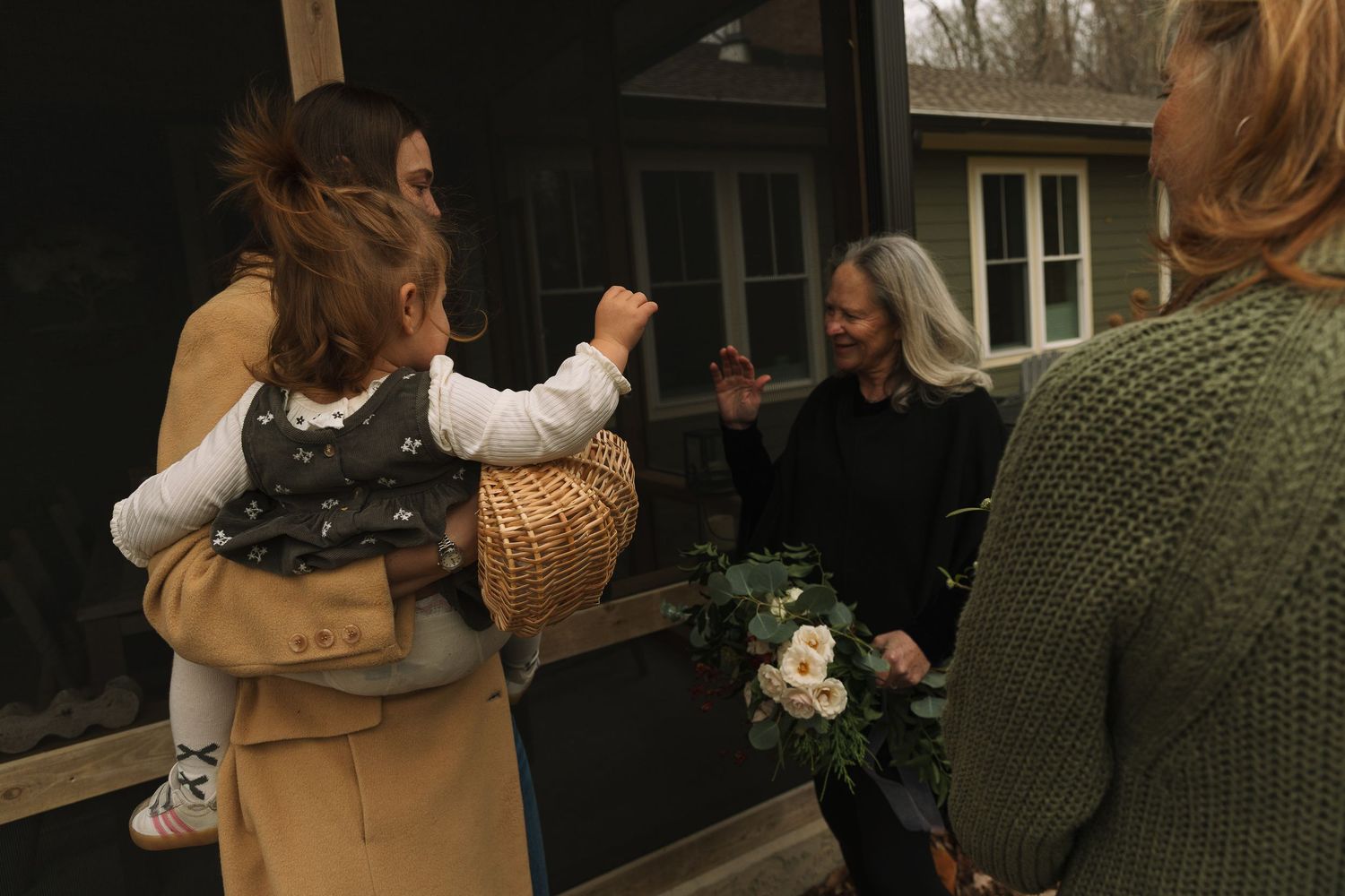 Women gathering outdoors, one holding a baby in a basket, greeting another woman holding white flowers.