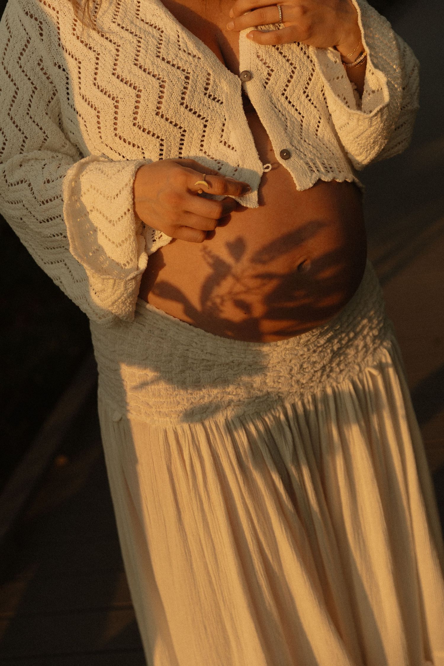 Pregnant woman in white crochet cardigan and skirt with floral shadow on baby bump.