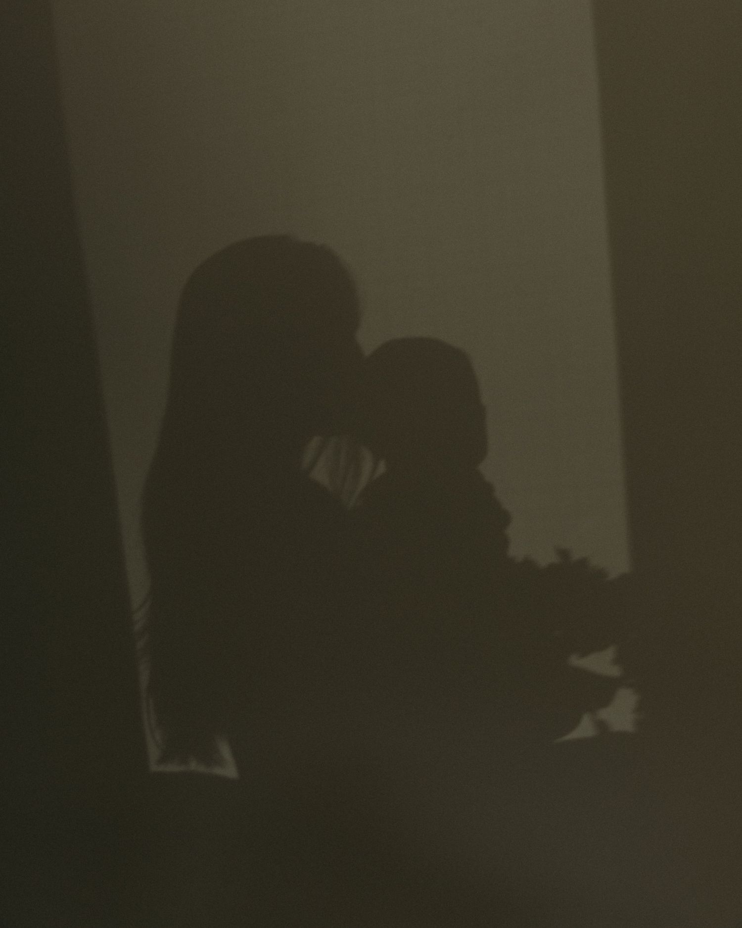 Silhouette of a mother tenderly holding her baby against a softly lit wall, creating a moody, intimate shadow portrait.