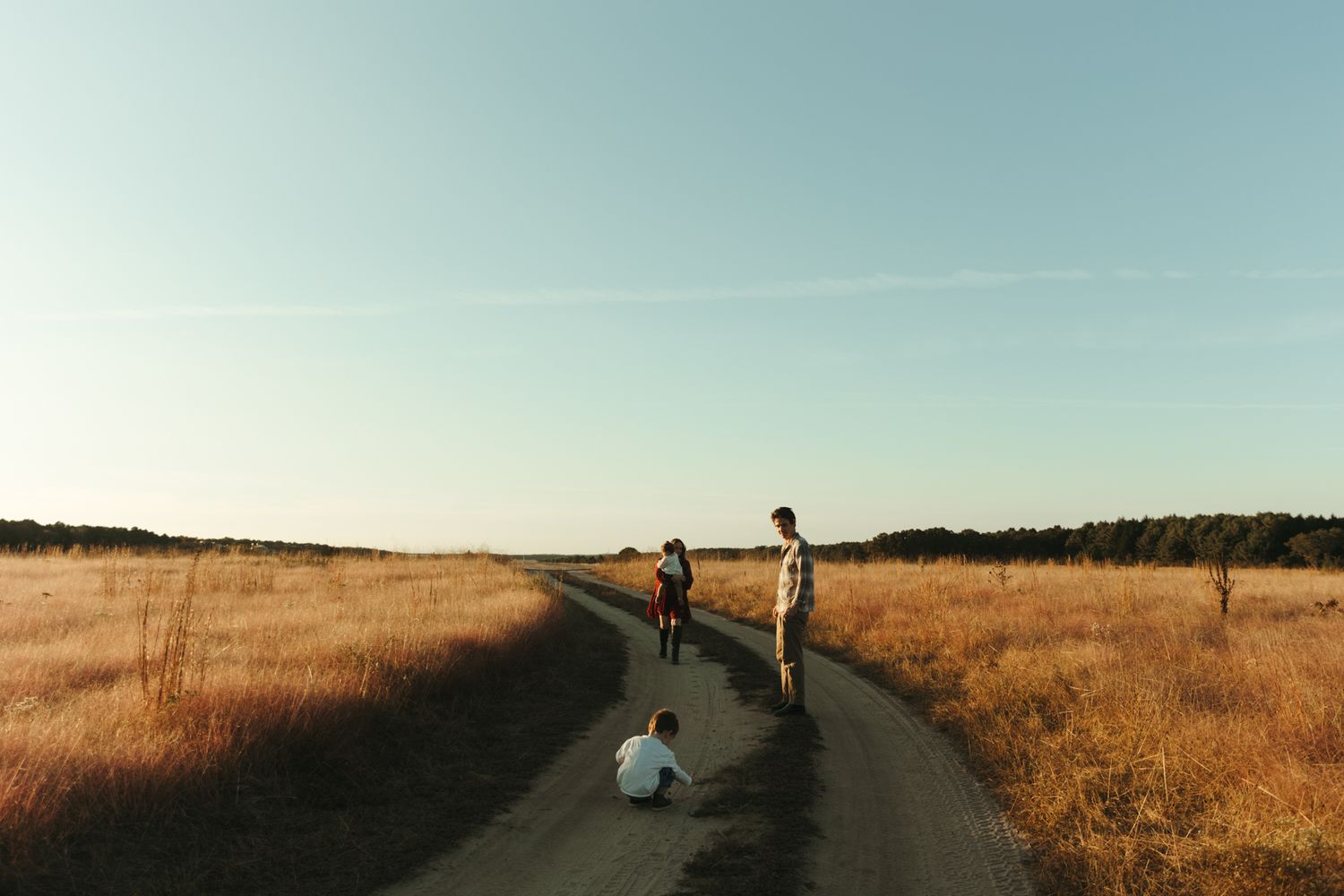 Family of three on a rural dirt road surrounded by golden fields under a clear blue sky.