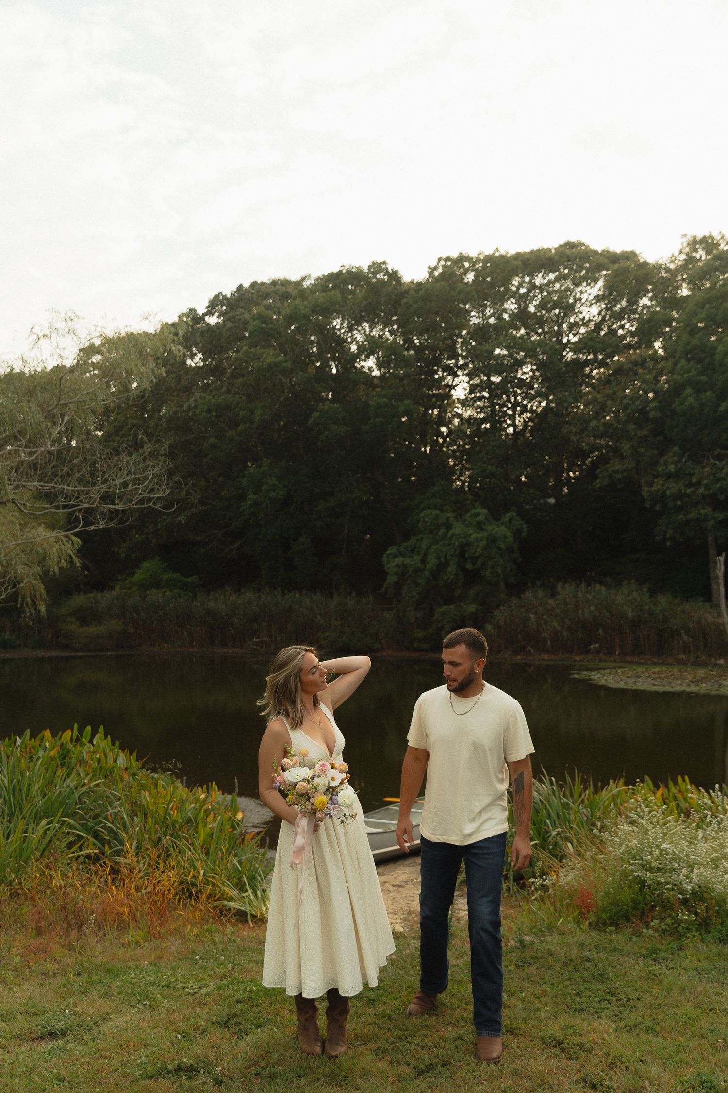 Engaged couple holding bouquet of wildflowers by serene pond surrounded by lush green trees.