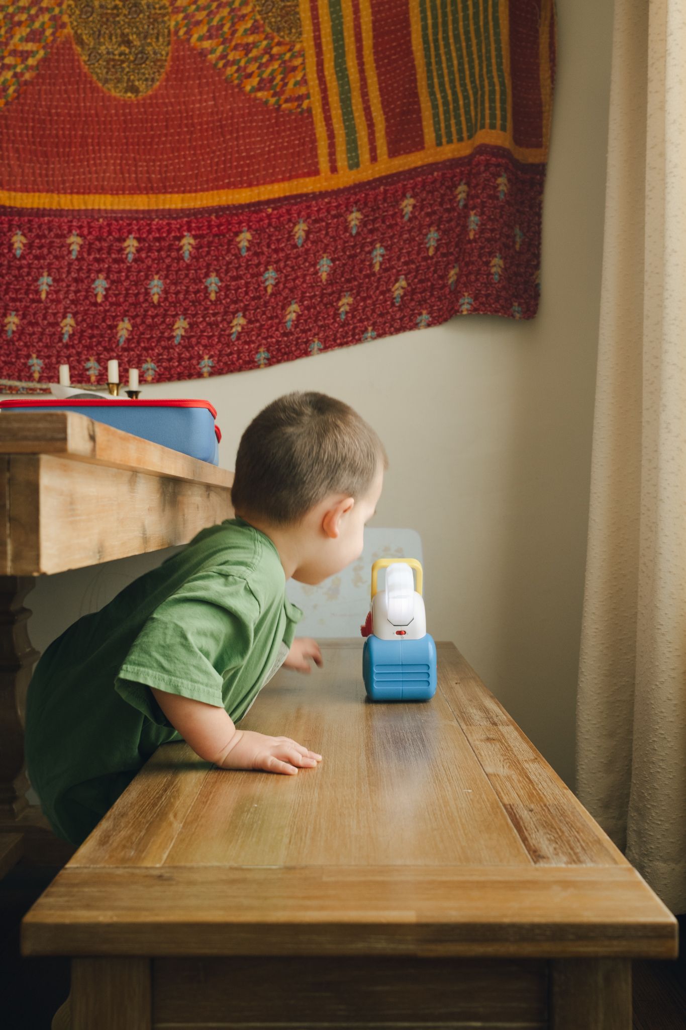Child in green shirt leans over wooden table to look at small blue and white toy on surface.