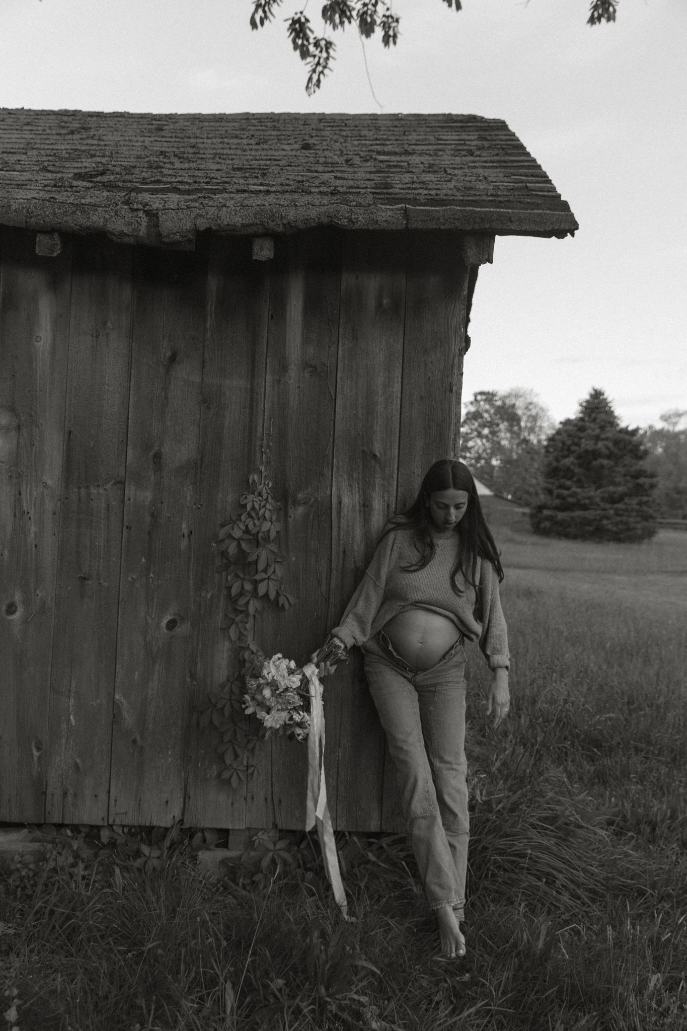 Black and white photo of pregnant woman holding flowers beside rustic wooden barn in rural field.