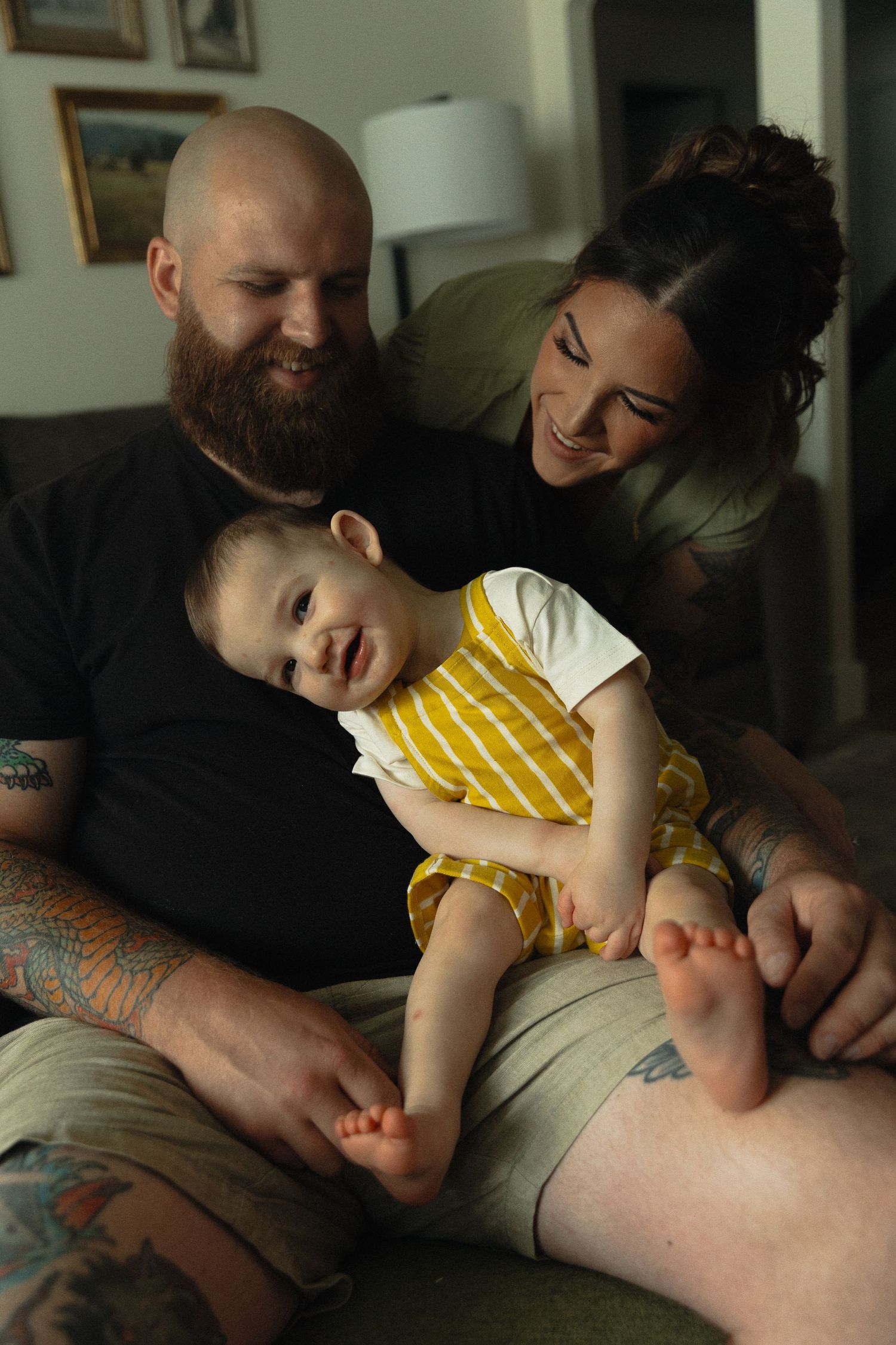 A tattooed bald man and dark-haired woman smile joyfully at a happy baby in a yellow outfit on a couch.