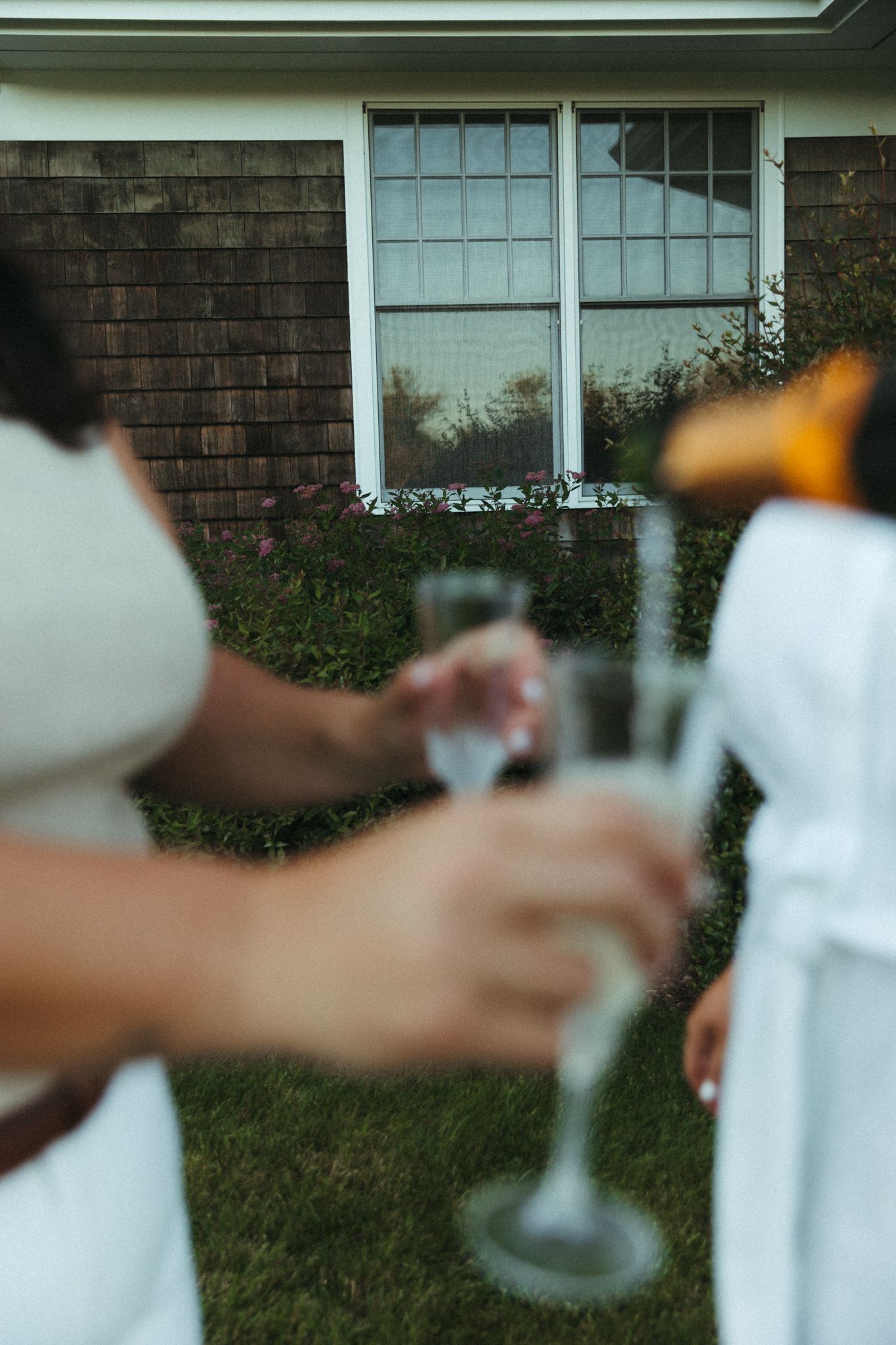Two people in white attire toasting with champagne flutes outdoors near a house with a flower window box.