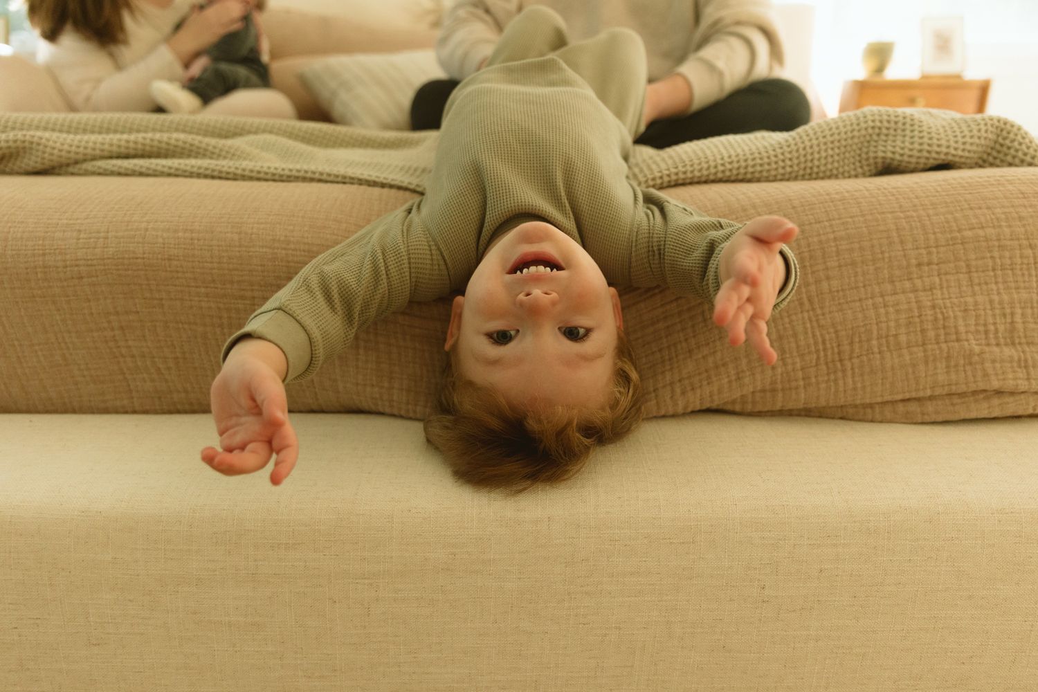 A playful young child hanging upside down off a beige sofa, smiling joyfully, with adults sitting in the background.