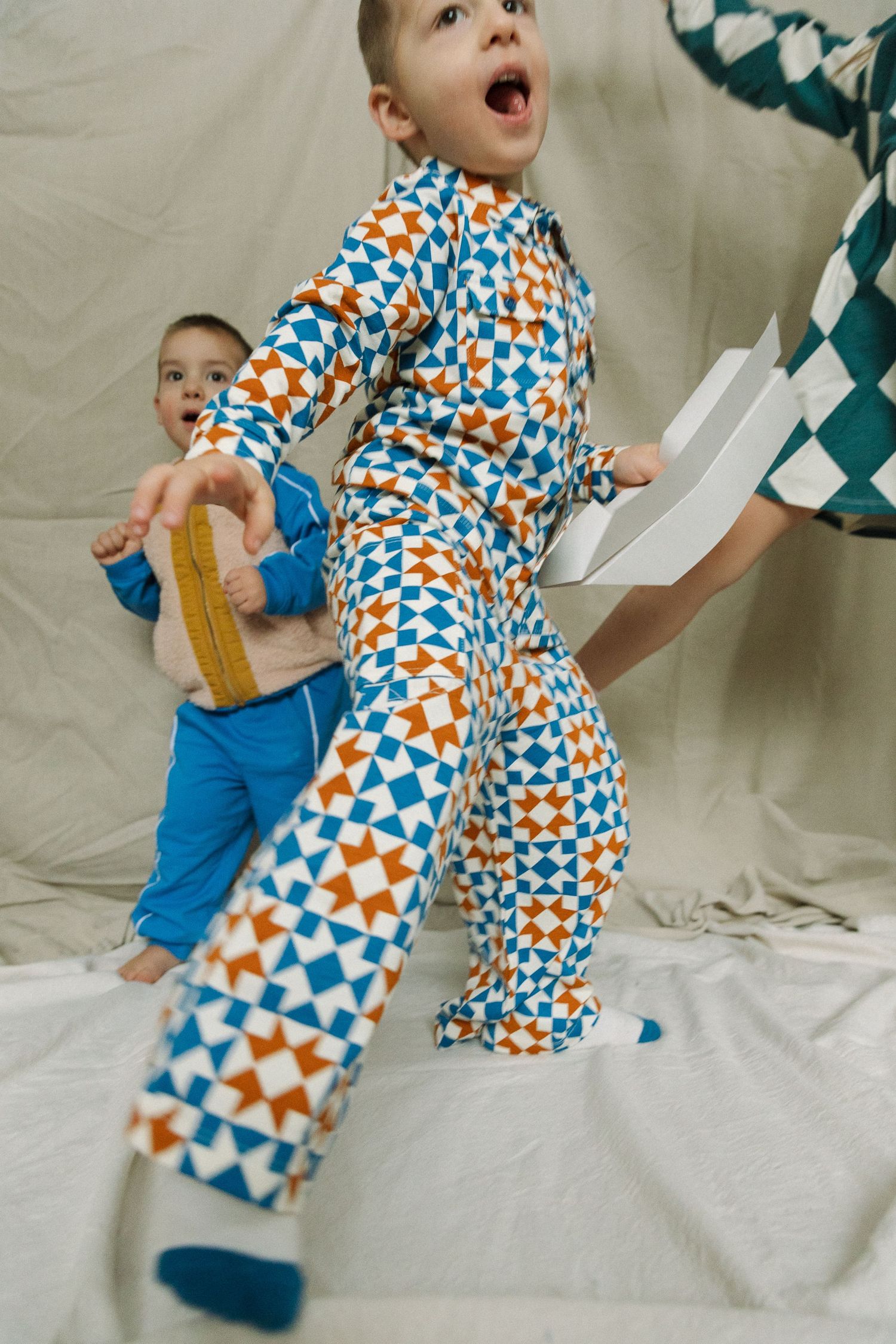 Two children playing energetically in colorful geometric patterned jumpsuits, one with mouth open mid-action.