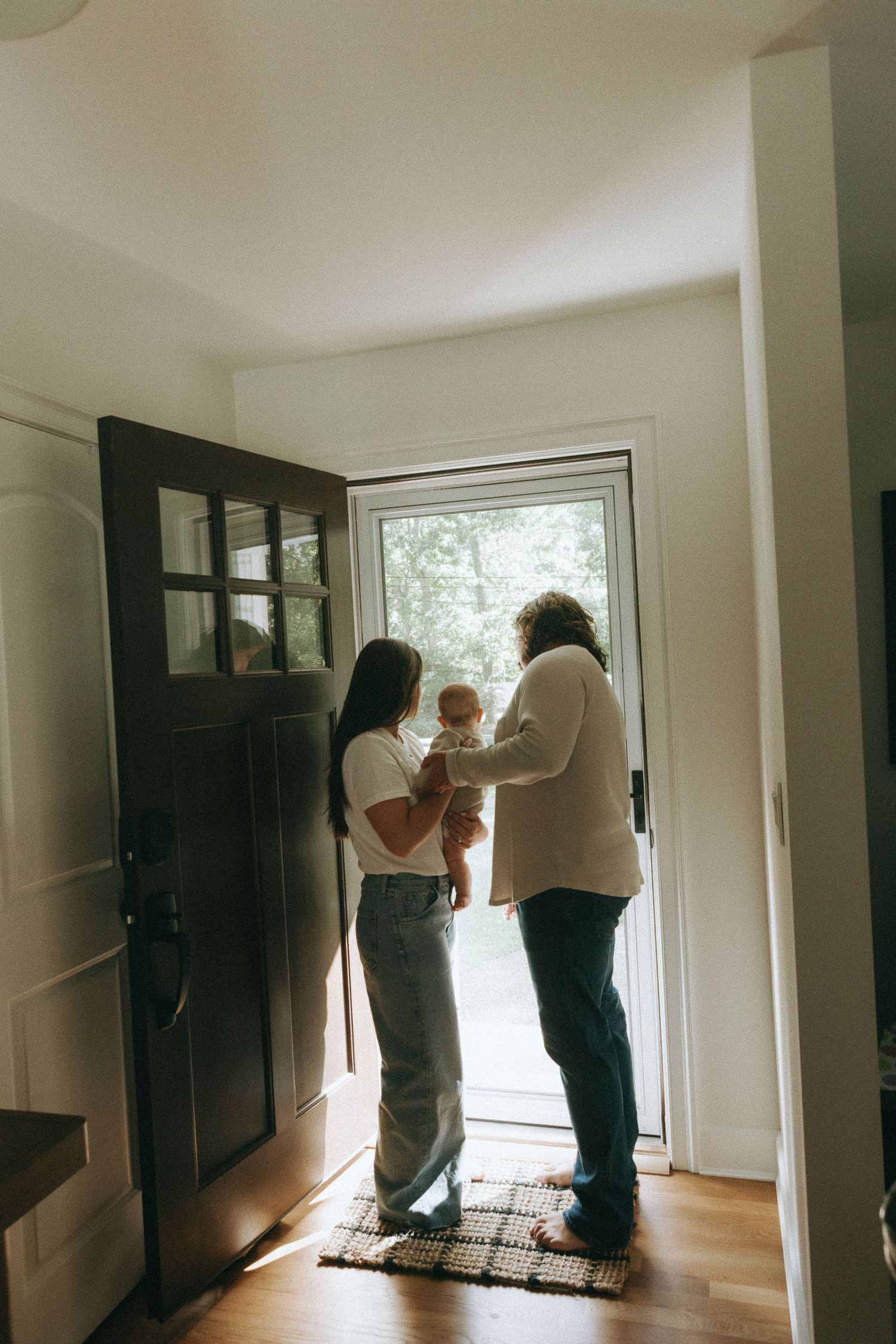 Family with newborn baby standing in a bright home entryway by an open front door.