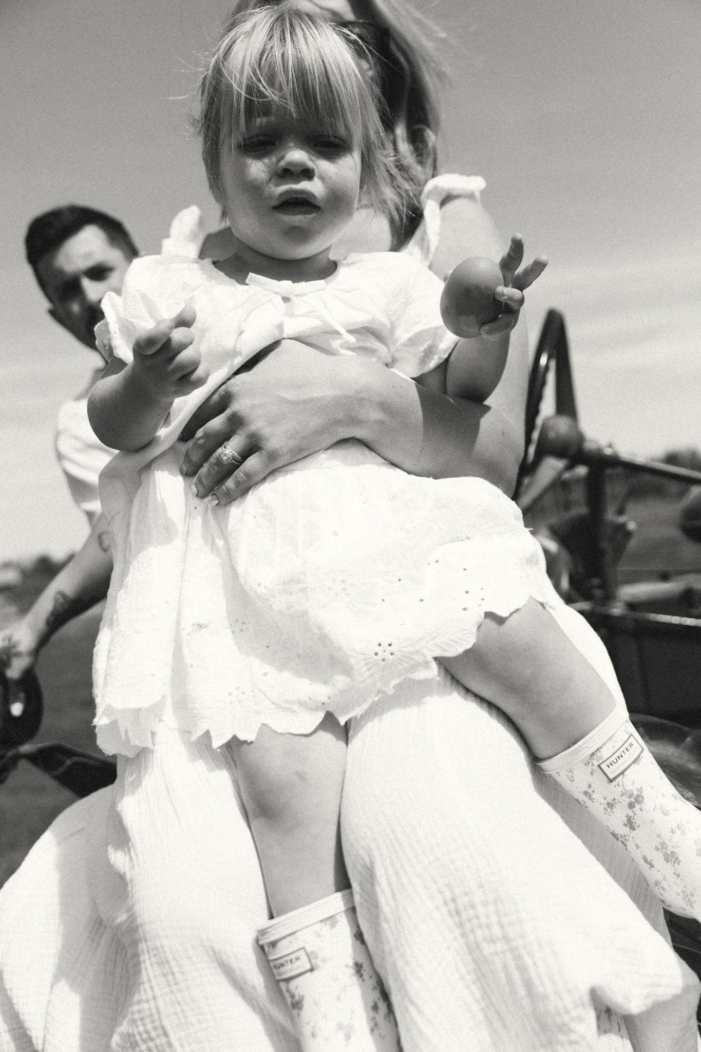 Black and white photo of a toddler girl in white dress being held by an adult outdoors near vintage machinery.