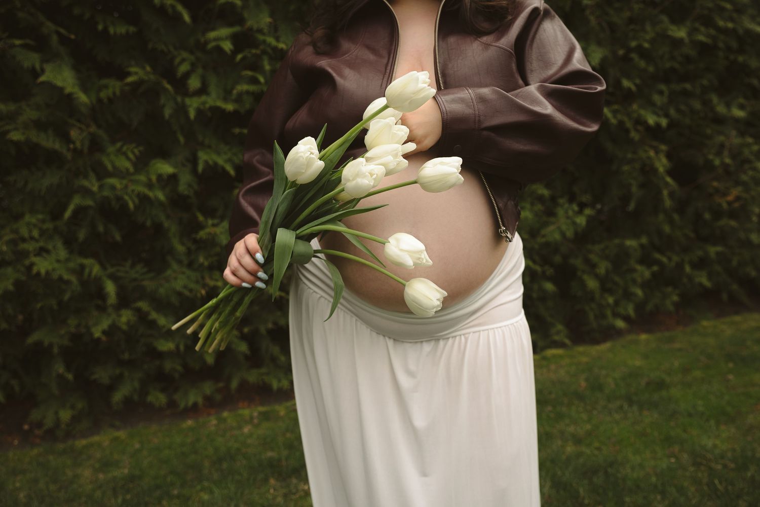 Pregnant woman in brown top and white skirt holding white tulips bouquet, showcasing baby bump outdoors.