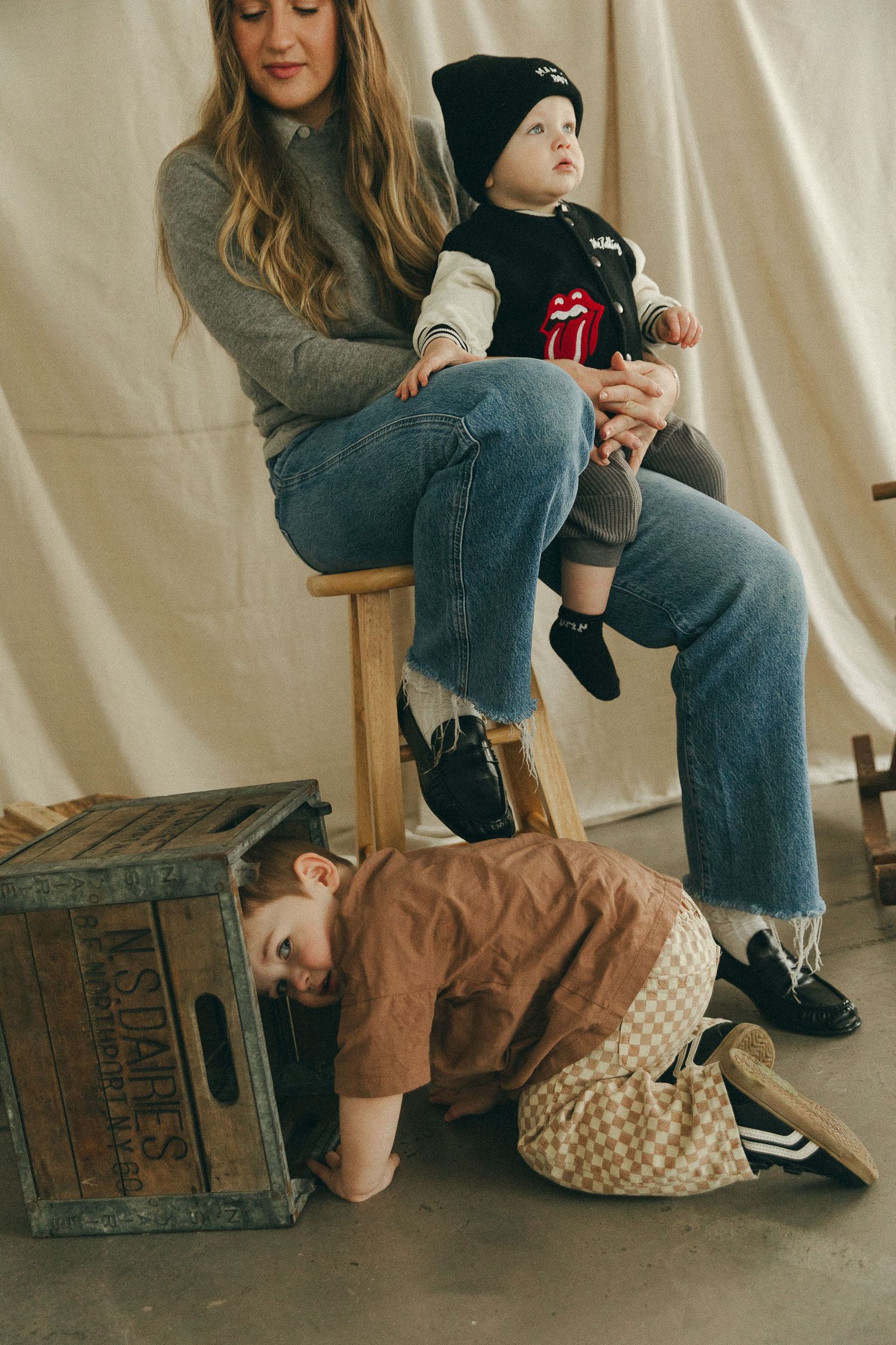 Woman sitting on stool holding baby in Rolling Stones shirt while toddler in brown jacket plays beneath her.