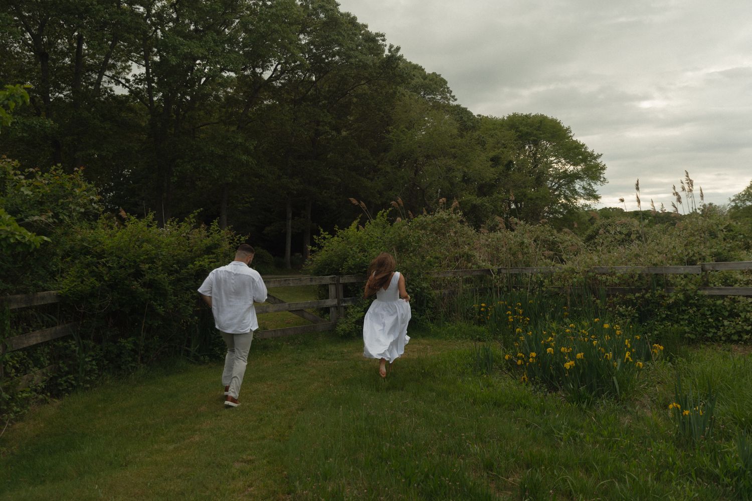 Two people in white clothing walk through a lush green garden path surrounded by trees and wildflowers.