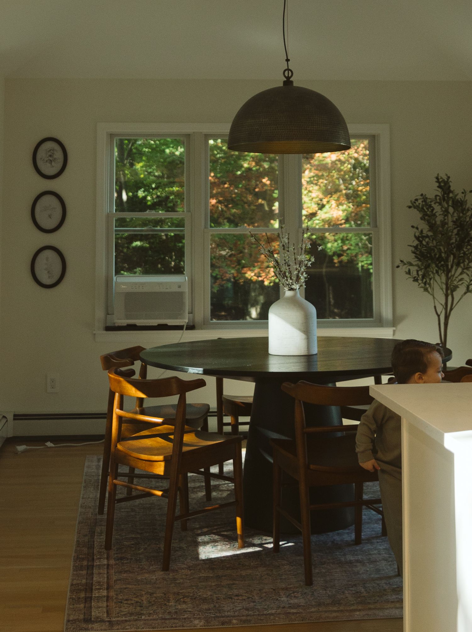 Cozy dining room with round dark wood table, woven chairs, dome pendant light, and large window overlooking lush green trees.