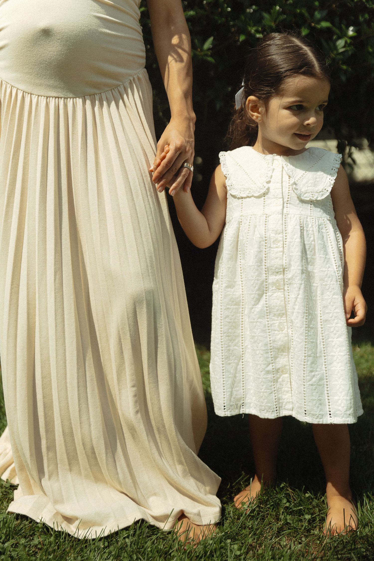 Young girl in white dress holding hands with a pregnant woman in a sunny garden.
