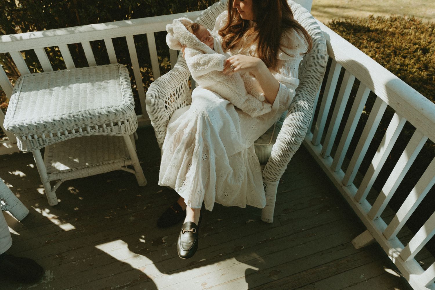 Woman in white dress sitting in wicker chair on wooden porch, cradling a baby in soft natural sunlight.