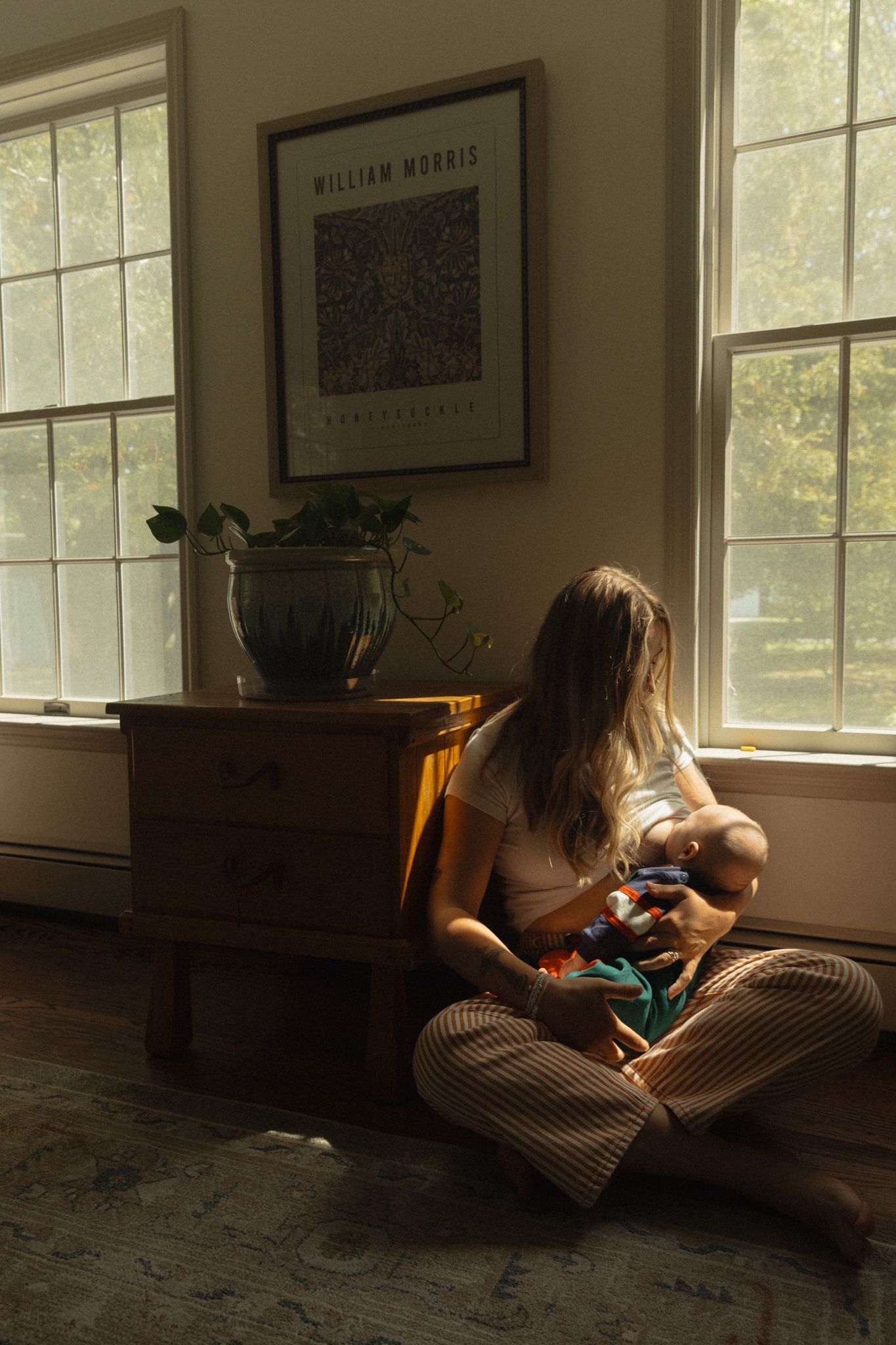 Mother breastfeeding newborn baby while sitting cross-legged on floor beside dresser in sunlit room.