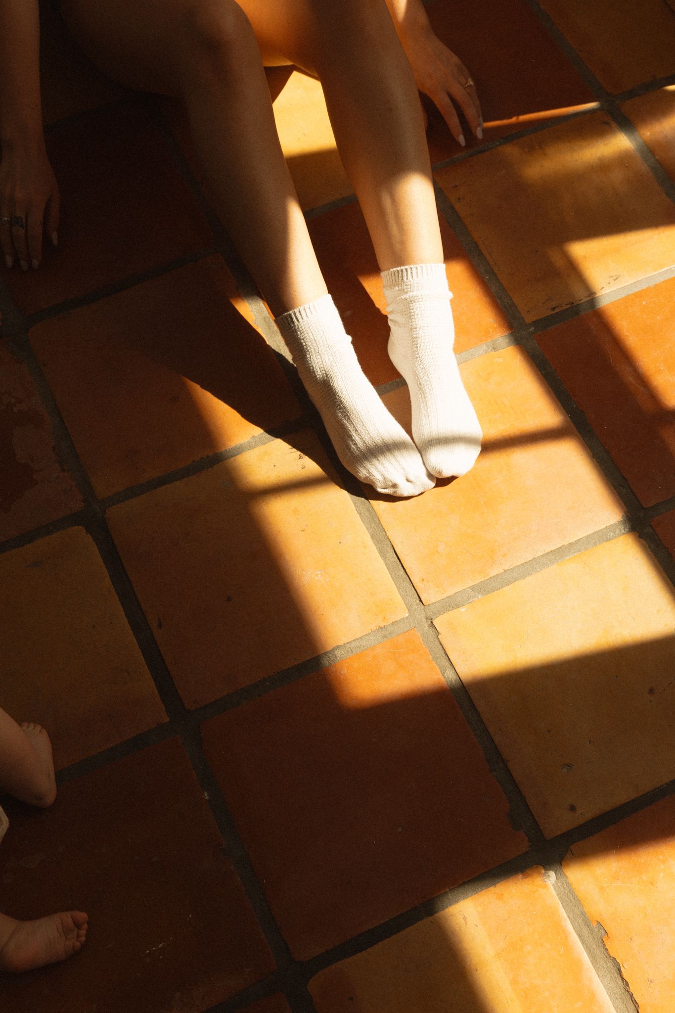 Person sitting on terracotta tile floor wearing white ankle socks in warm sunlight with dramatic shadows.