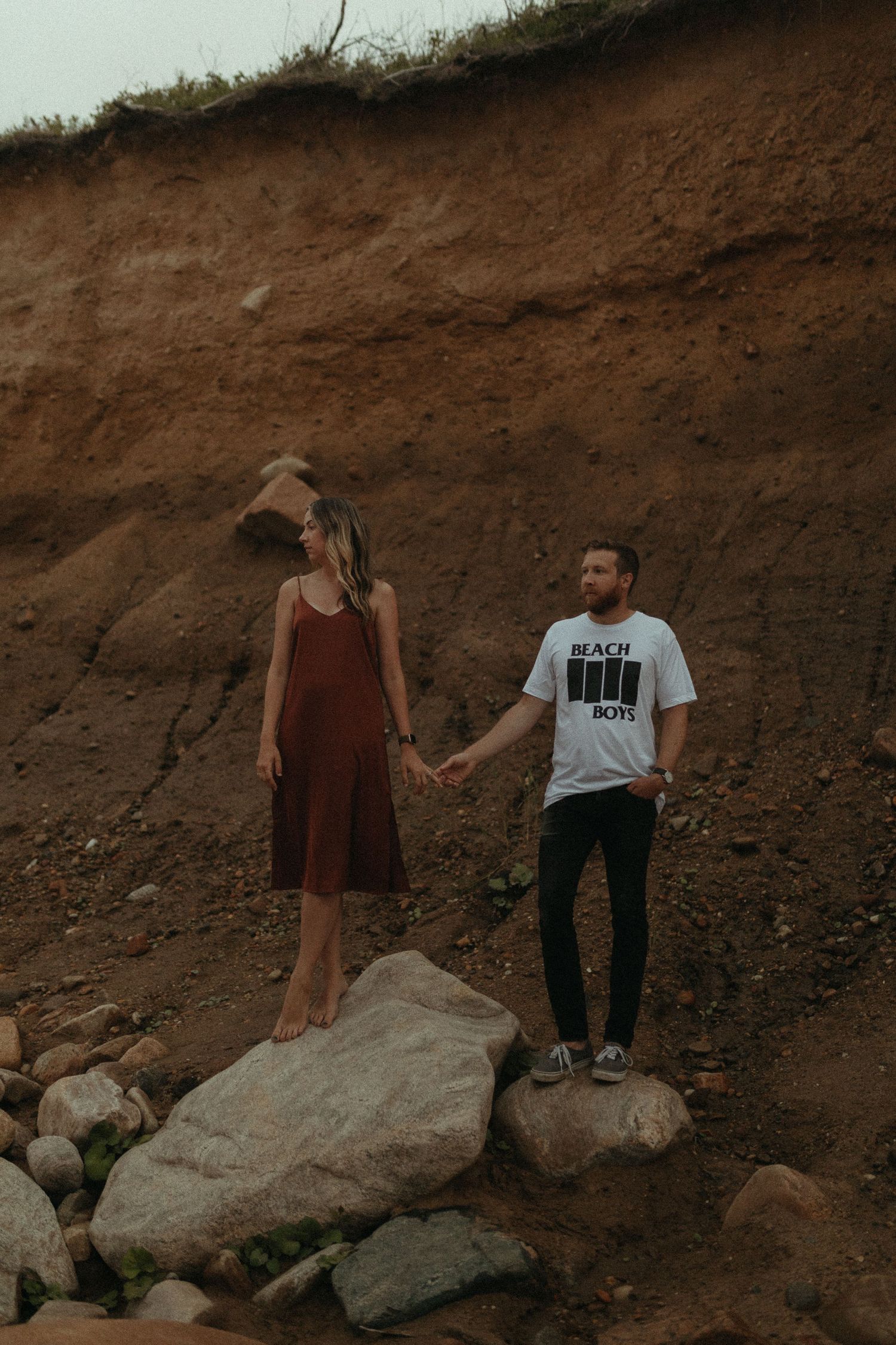 Couple holding hands on rocky terrain in front of an earthy cliff, man wearing Beach Boys tee.