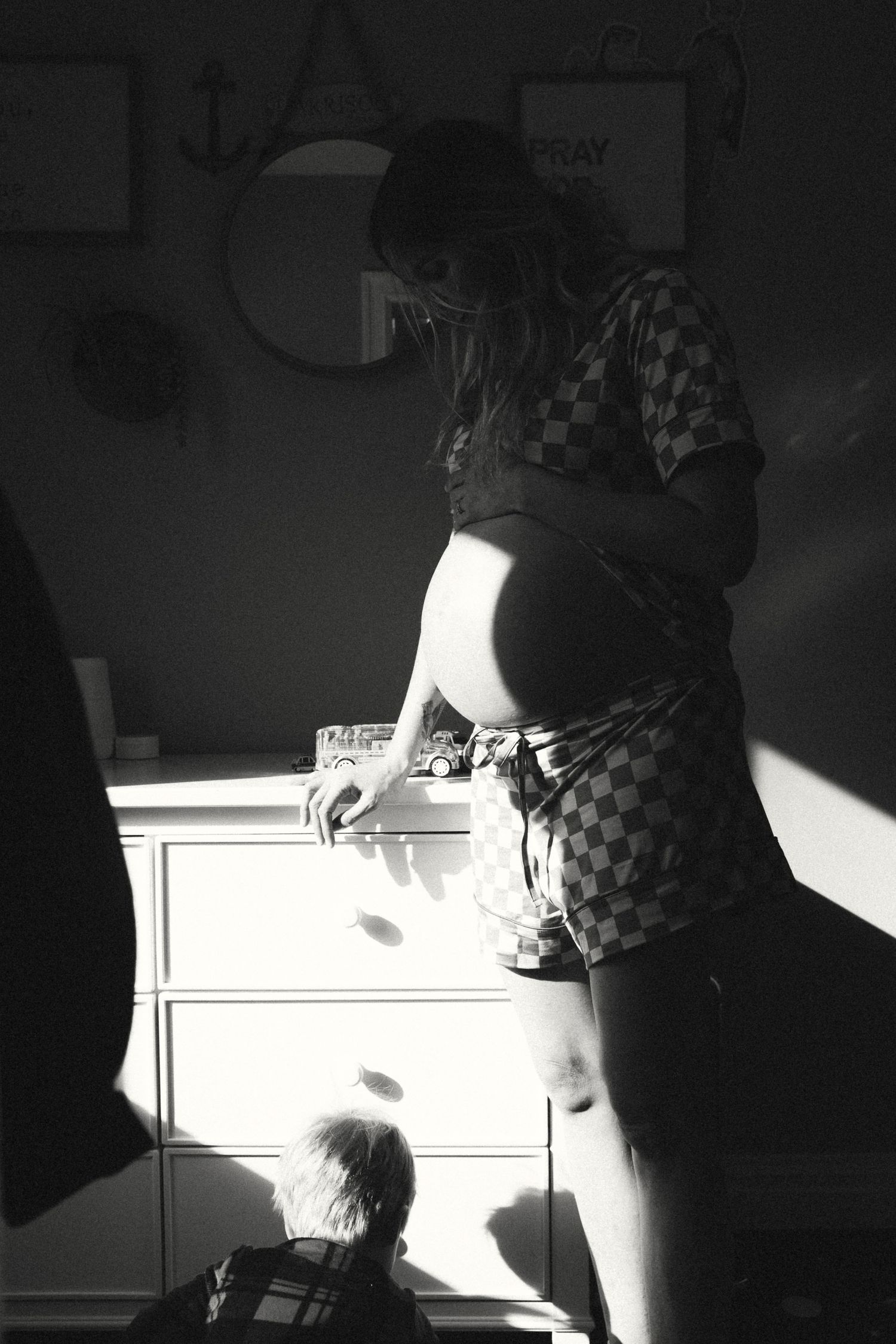 A dramatic black and white photo of a pregnant woman standing by a dresser with a child nearby in natural light.