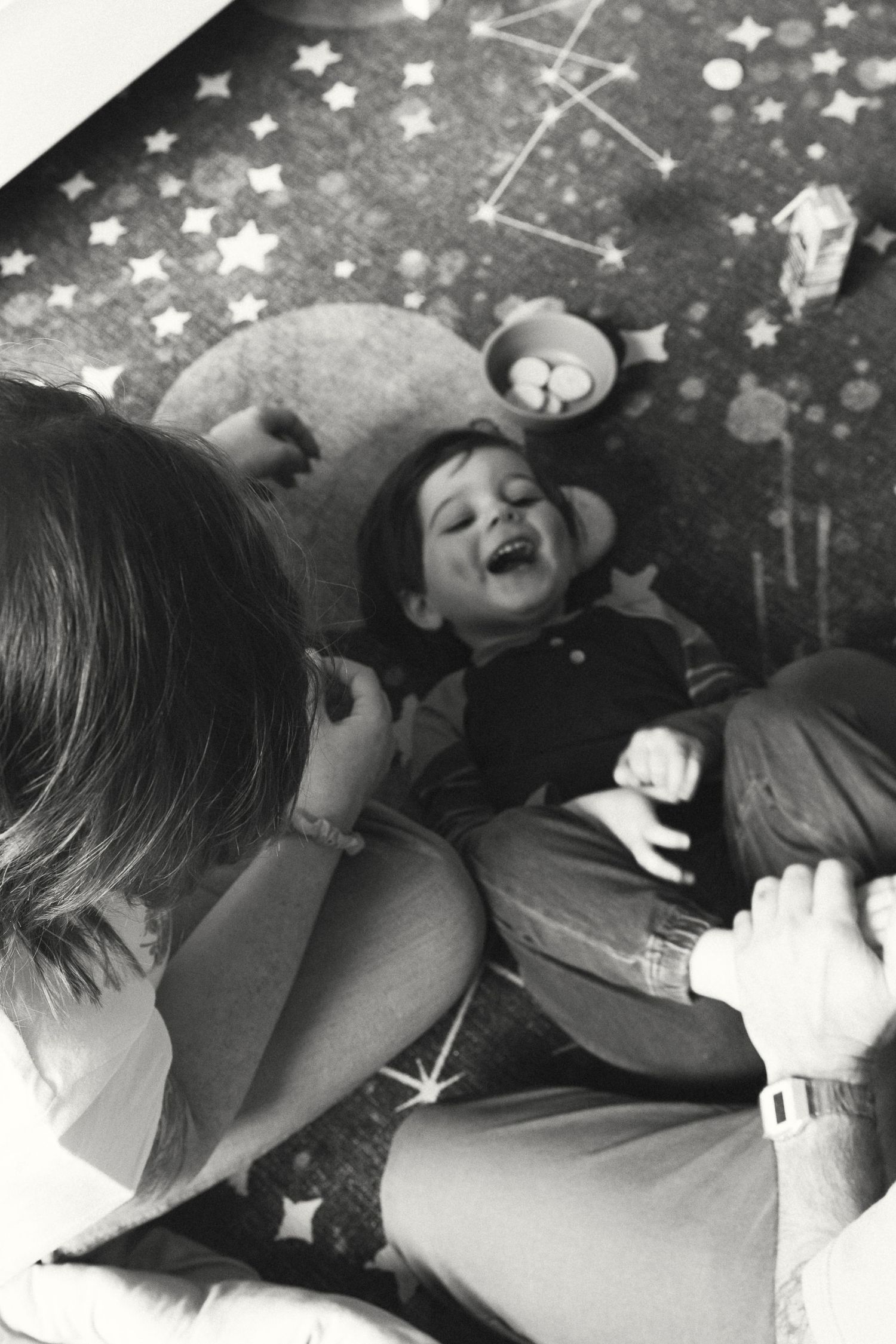 Black and white photo of a laughing toddler lying on a star-patterned rug, surrounded by adults.