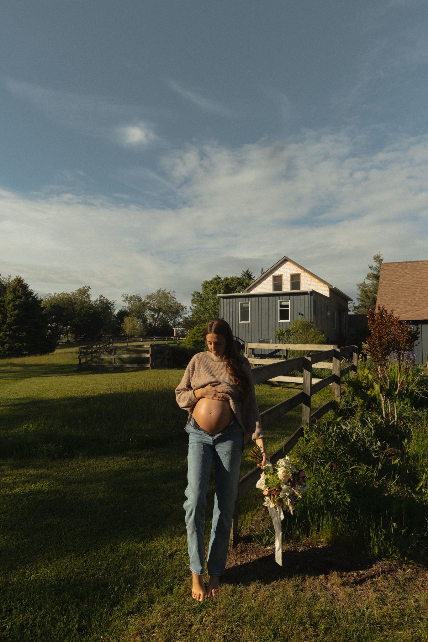 Pregnant woman in jeans holding flowers stands in a sunny farm yard with a gray barn and wooden fence.