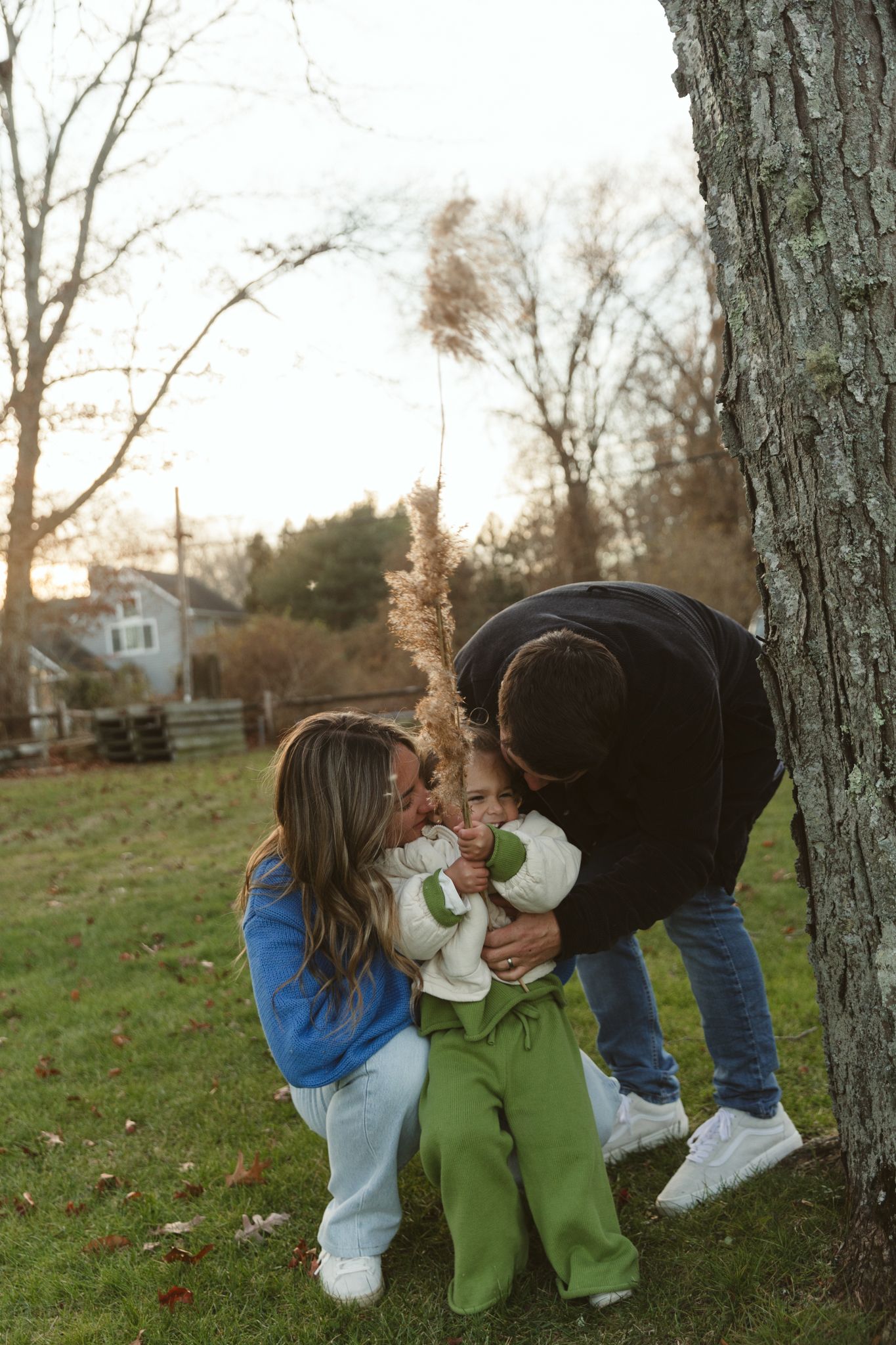 Parents lovingly embracing toddler in green outfit near large tree trunk at golden hour.