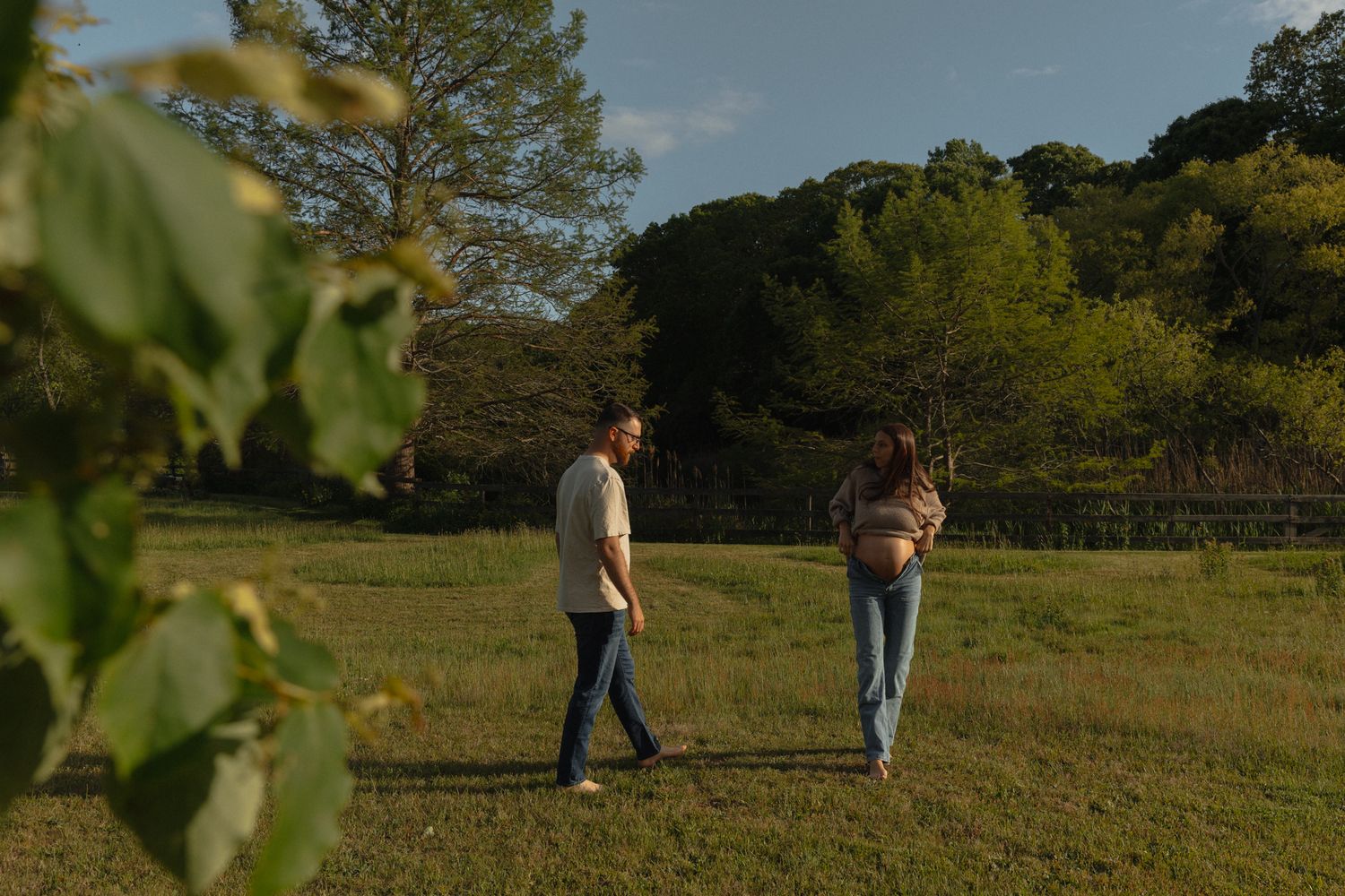 Expectant couple walking together through green meadow on sunny day surrounded by trees.