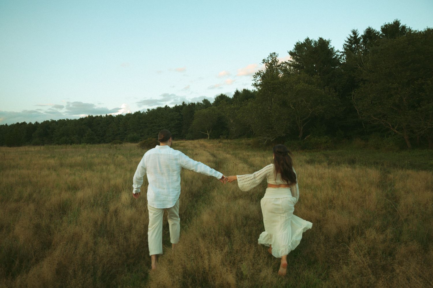 Couple holding hands walking toward forest through golden grass field at dusk.
