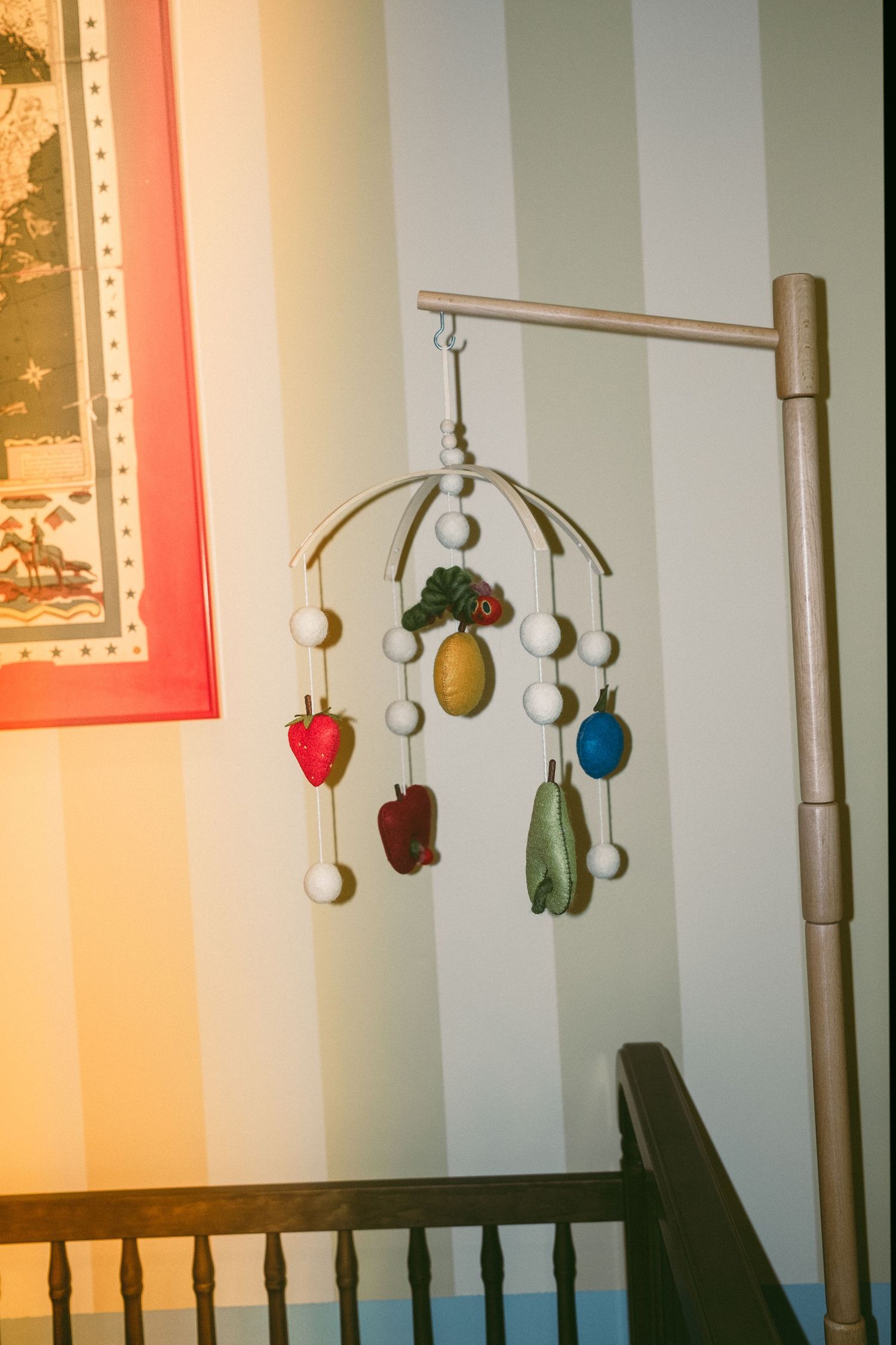 A colorful vintage baby mobile with fruit shapes hangs over a wooden crib in a striped nursery.