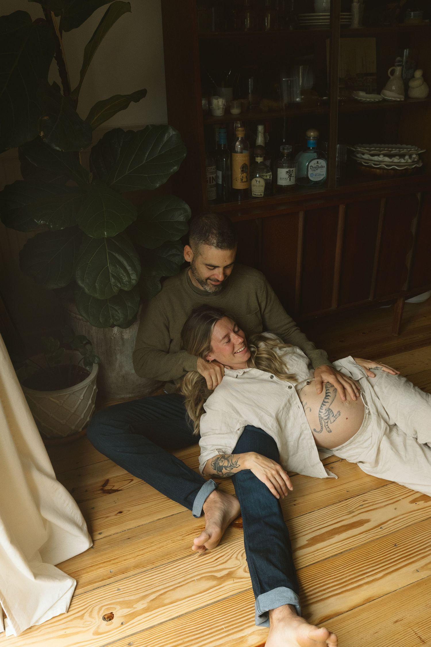 Couple relaxing on wooden floor, pregnant woman lying on man's lap, tattoos visible, cozy home interior with plants.
